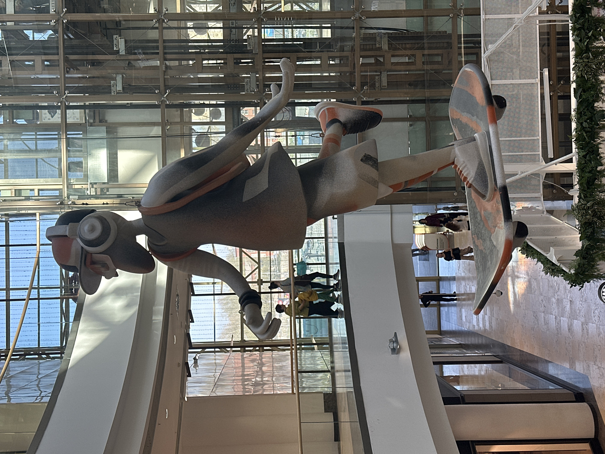 Here is a description of the image:
The image depicts a large sculpture of a skateboarder suspended in the air within a modern, multi-story building with a glass facade and exposed architectural elements. The sculpture is stylized and appears to be made of a composite material, with a color palette of grey, orange, black, and white. The figure wears headphones, a wristband, and shoes with orange accents. The skateboard is multi-colored with orange and gray stripes and black wheels. The building's interior features metal and glass structures, creating a grid-like pattern in the background. People can be seen reflected in the glass surfaces. Greenery hangs from the top-right corner of the frame. The sculpture appears to be in Novosibirsk, Russia.