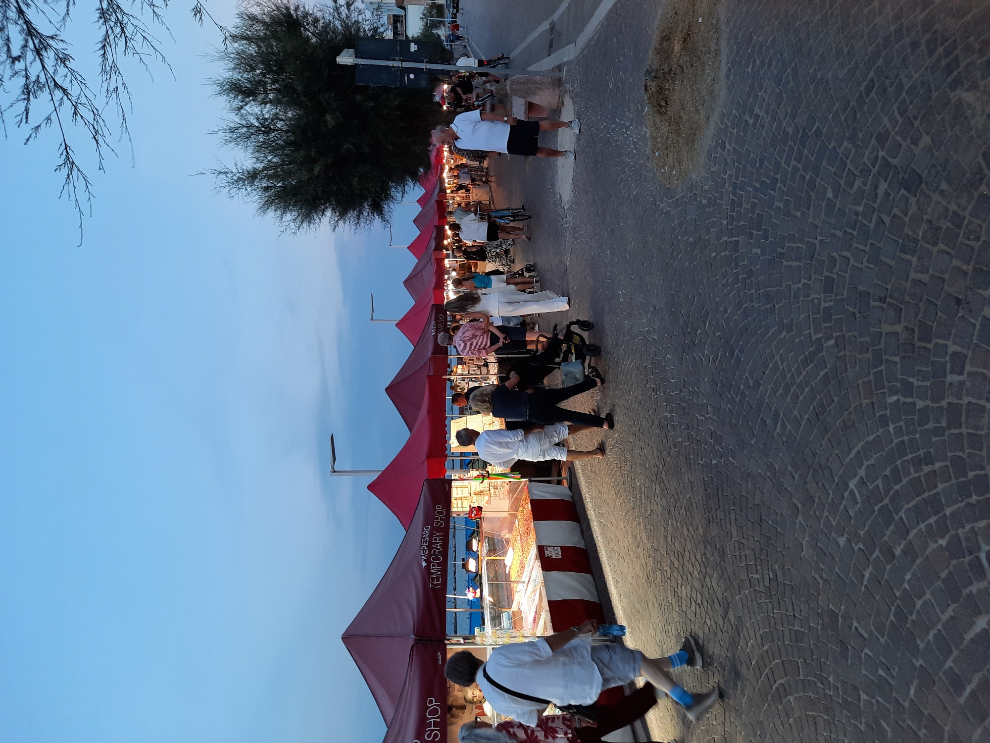 The image depicts a lively outdoor market scene at dusk on a cobblestone promenade in Pesaro, Italy. Numerous red tents, identified by the text "Wepesaro Temporary Shop," line the walkway, suggesting a commercial event. People are seen browsing the stalls, walking, and conversing. Some tents display products, including what appears to be food items in glass cases. The promenade runs alongside the sea, with glimpses of water visible between the tents. The sky is a gradient of pale blue and twilight hues. Streetlights are visible, and the overall atmosphere is one of casual activity and commerce.