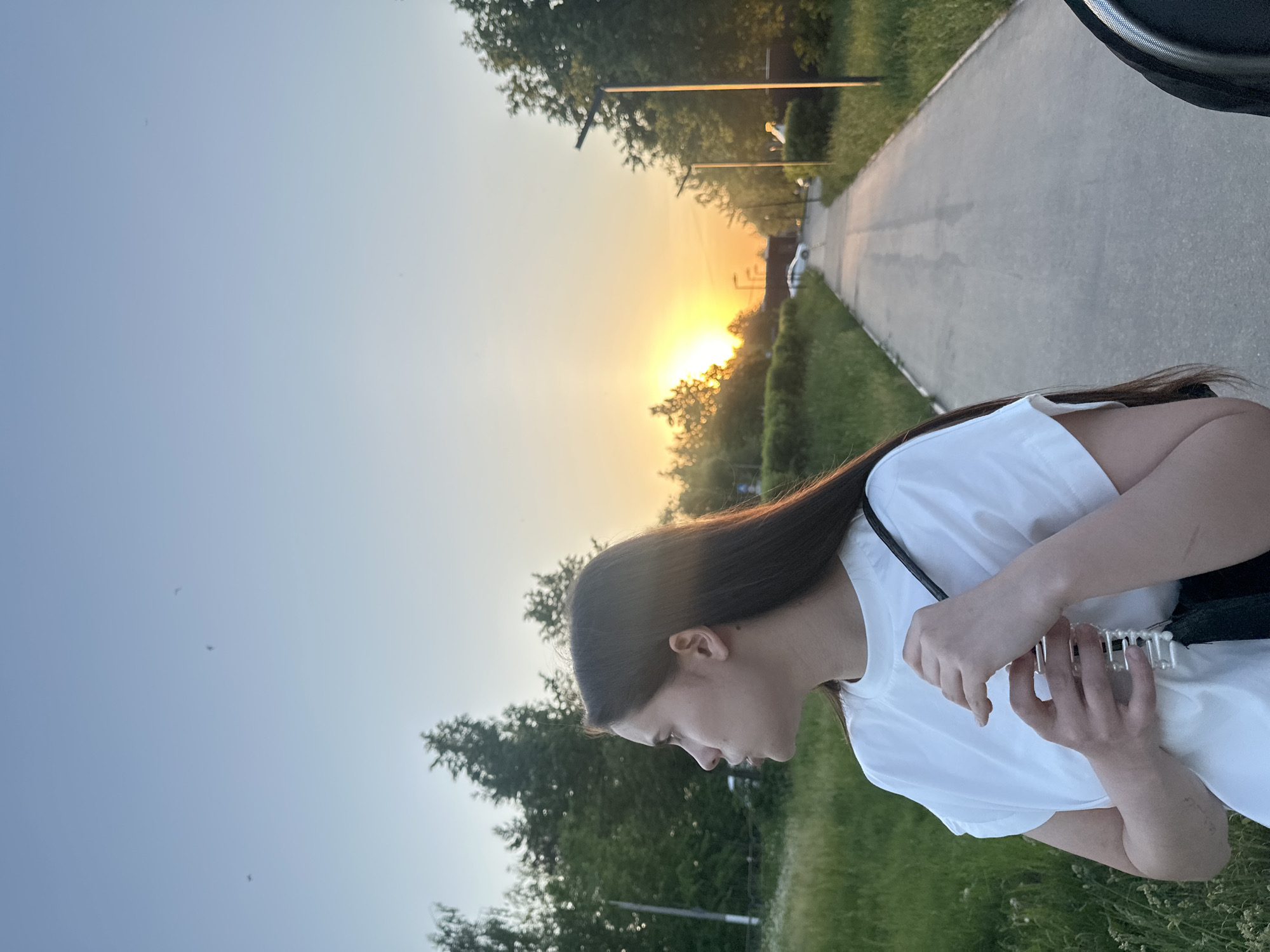 A vertical, medium shot photograph taken outside during sunset, showing a young woman in the foreground and a paved road receding into the distance.
The woman is turned to her left, with her head tilted down, as if looking at something she's holding. She has long, dark hair and is wearing a white t-shirt with a black strap visible on her shoulder. Her hands are holding a translucent object with white pearl-like accents.
The road stretches into the distance, lined with trees and greenery. The setting sun creates a bright glow between the trees and casts a warm light on the scene. Streetlights are visible along the road, and a few vehicles can be seen in the far distance.
The environment suggests a park or recreational area. Given the location is Chaikovsky, Russia, this might be a park near the city's infrastructure, close to a roadway.