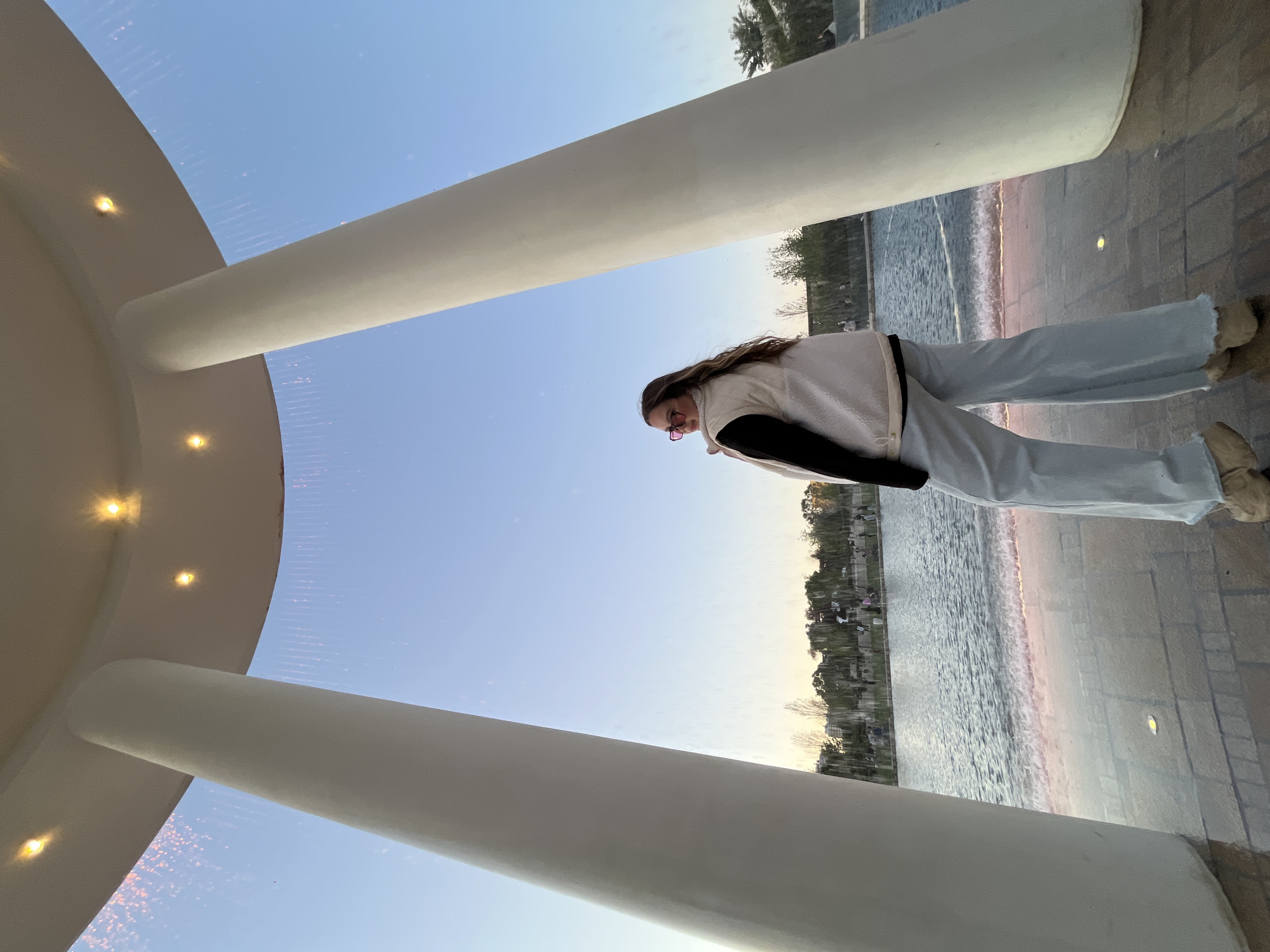 A rotated, eye-level shot depicts a woman standing beneath a large, circular structure supported by white columns. Small lights dot the interior of the structure. The woman is wearing light-colored jeans, a white jacket with a black shirt underneath, and glasses. Outside the structure, a water feature or fountain is visible, reflecting the surrounding trees and buildings. The scene is set against a light blue sky, indicating either early morning or late afternoon. Given the context of Krasnodar, Russia, the location is likely the Musical Fountain.