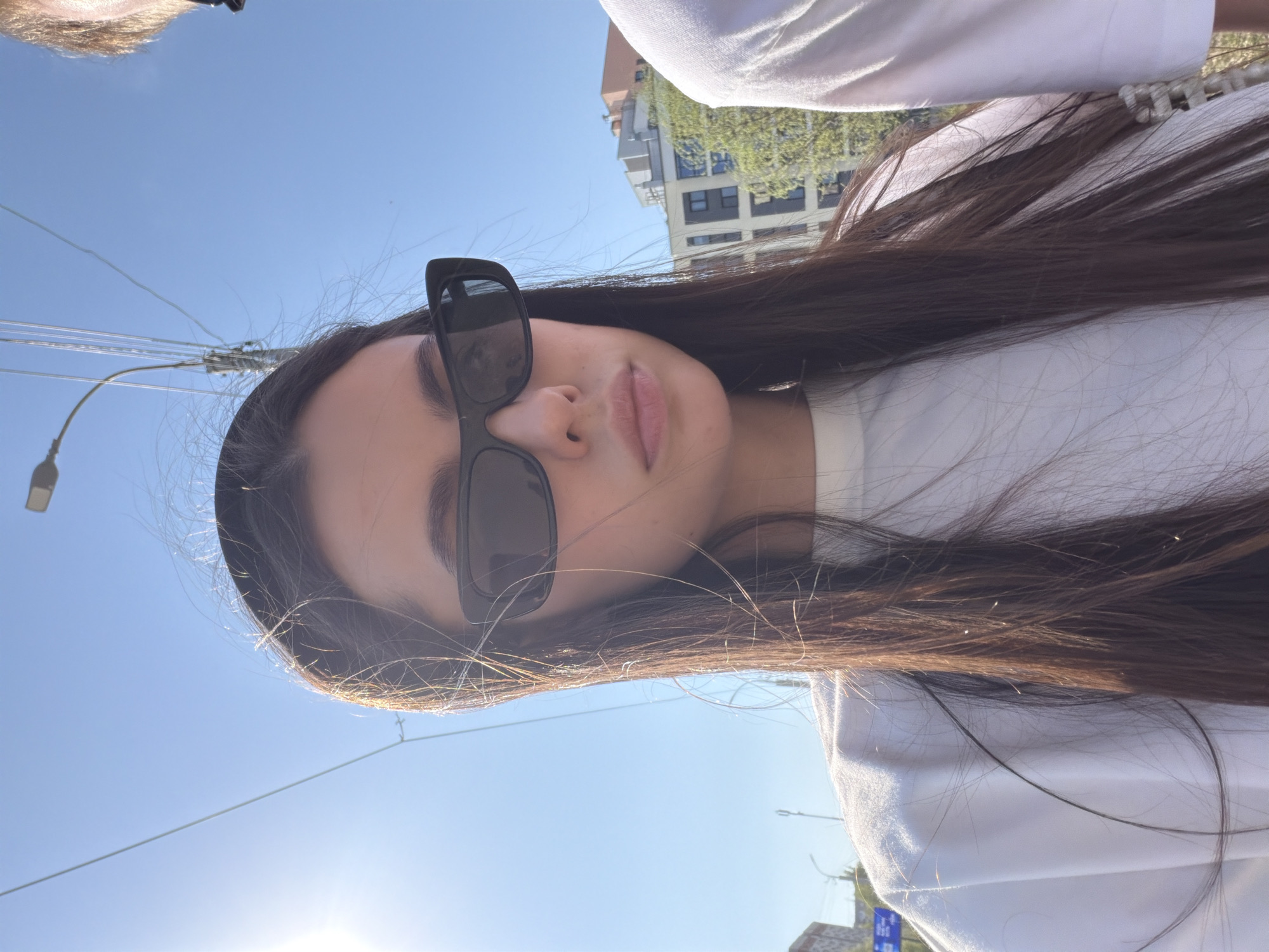 A young woman with long brown hair and sunglasses poses in an outdoor setting, likely in Izhevsk, Russia. Buildings are visible in the background. Overhead power lines are present in the composition. She is wearing a white shirt. The photo is taken from a low angle, looking up. The sky is clear and blue.
