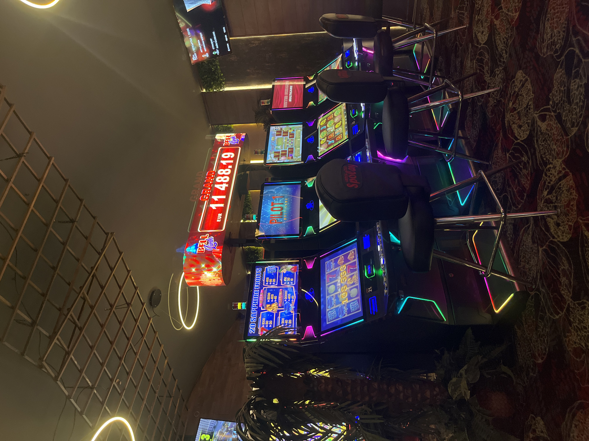 The image depicts a row of slot machines in what appears to be a casino or gaming establishment. The machines are brightly lit with colorful displays and neon lights, and they are equipped with chairs. A large sign above the machines displays a jackpot amount of "EUR 11,488.19". The casino has a modern design with decorative lighting, including circular fixtures hanging from the ceiling. There's greenery at the base of the machines and a patterned carpet on the floor. There are also television screens in the background, one of which displays an Aston Villa vs. Nottingham Forest football match. The image also includes text in Bulgarian, indicating that the scene is likely located in Bulgaria. There's no specific information suggesting the location to be in the city of Veliko Tarnovo.