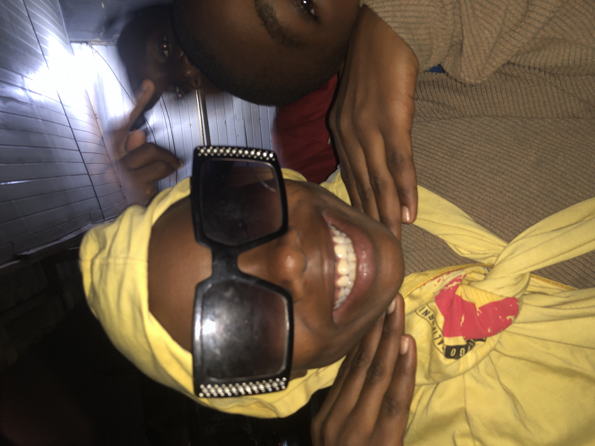 In the city of Ado, Nigeria, an individual, possibly a young person, is prominently featured in a close-up photograph. They are wearing a bright yellow hooded garment, and their face is adorned with large, black-rimmed sunglasses, the frames of which are embellished with small, reflective stones. The subject is smiling, revealing their teeth, and their hands are positioned around their neck and chin, with one hand resting on the yellow hood. Behind them, two other individuals are partially visible. One person is further back, with their face obscured by shadows, but their hand is raised, with fingers extended in a gesture. Another individual is to the right, with their head tilted and a portion of their face and eye visible. The background appears to be an interior space with a corrugated metal ceiling, illuminated by a bright light source from the top right. The overall ambiance is informal and candid.