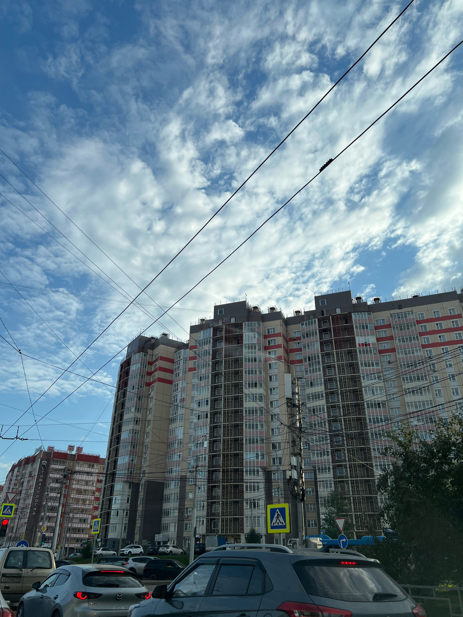 The image shows a view of a street in Krasnoyarsk, Russia, with a modern, multi-story residential building dominating the background. The building has a segmented facade with alternating panels of light-colored material, possibly concrete or composite, and large glass windows with dark frames. There are bands of red accents on some sections of the building. Several power lines and cables crisscross the sky in the foreground, obscuring parts of the building and the sky. The sky itself is partly cloudy with a mix of white cumulus clouds against a blue backdrop.
In the foreground, several cars are visible on a street, suggesting traffic. One car in the immediate foreground is dark gray, and another silver car is in front of it. Pedestrian crossing signs are visible on poles, one with a yellow background and a depiction of a person walking, and another with a blue background and a circular arrow indicating a turn. A traffic light showing red is also present. To the left of the main building, another similar residential building is partially visible. Trees and foliage are present on the right side of the image, partially obscuring the lower portion of the residential building. The overall scene depicts an urban environment with significant infrastructure, including high-rise buildings and overhead electrical lines.