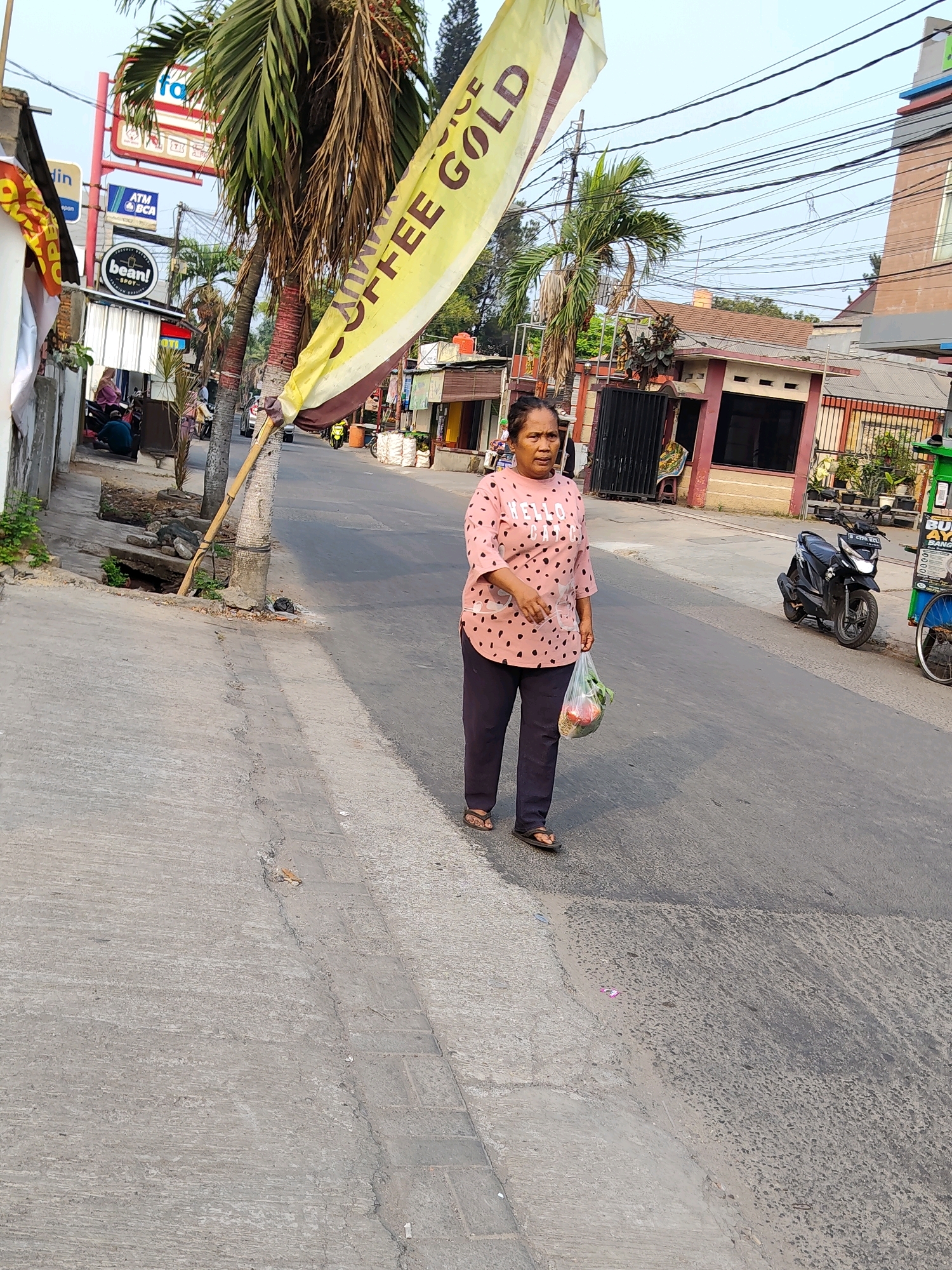 An overhead shot captures a woman in a pink polka-dot shirt and dark pants walking on a paved street in Bekasi, Indonesia. She carries a clear plastic bag filled with produce. Behind her, a motorcycle is parked on the side of the road, and a yellow banner with the text "COFFEE GOLD" is draped across a palm tree. Several buildings, including one with a red and white facade, line the street. The sky is visible above the buildings, with a network of electrical wires crisscrossing the scene. A concrete sidewalk runs alongside the street, and a drain opening is visible near the base of the palm tree.