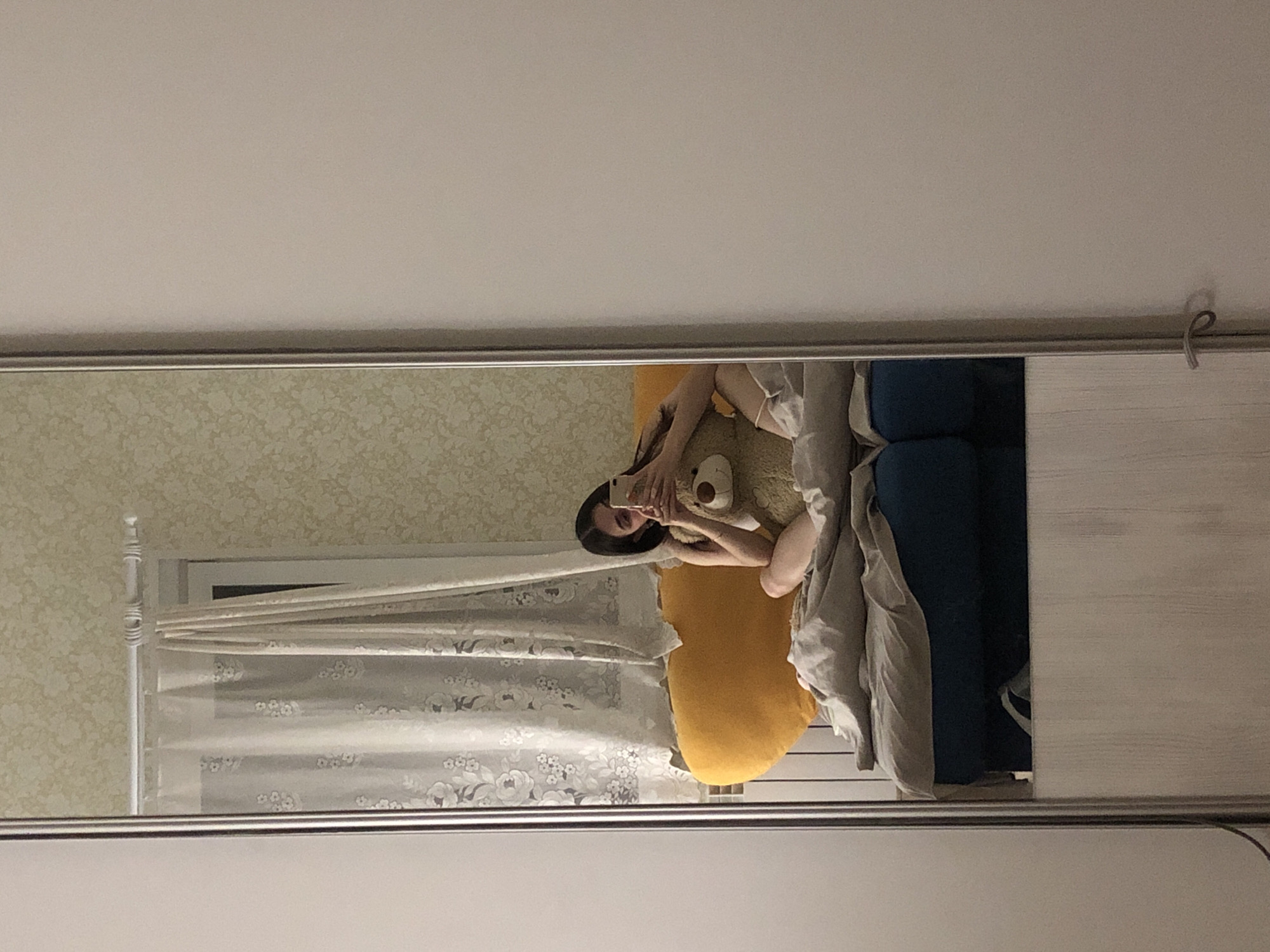 A person is taking a selfie in front of a mirror. They are holding a phone and are positioned on a yellow pillow. Beside the pillow is a blue object and a grey blanket. The mirror reflects a window with patterned curtains, a patterned wall, and a white closet door with a handle. There is no information about the location in the city of Noviy, Russia.
