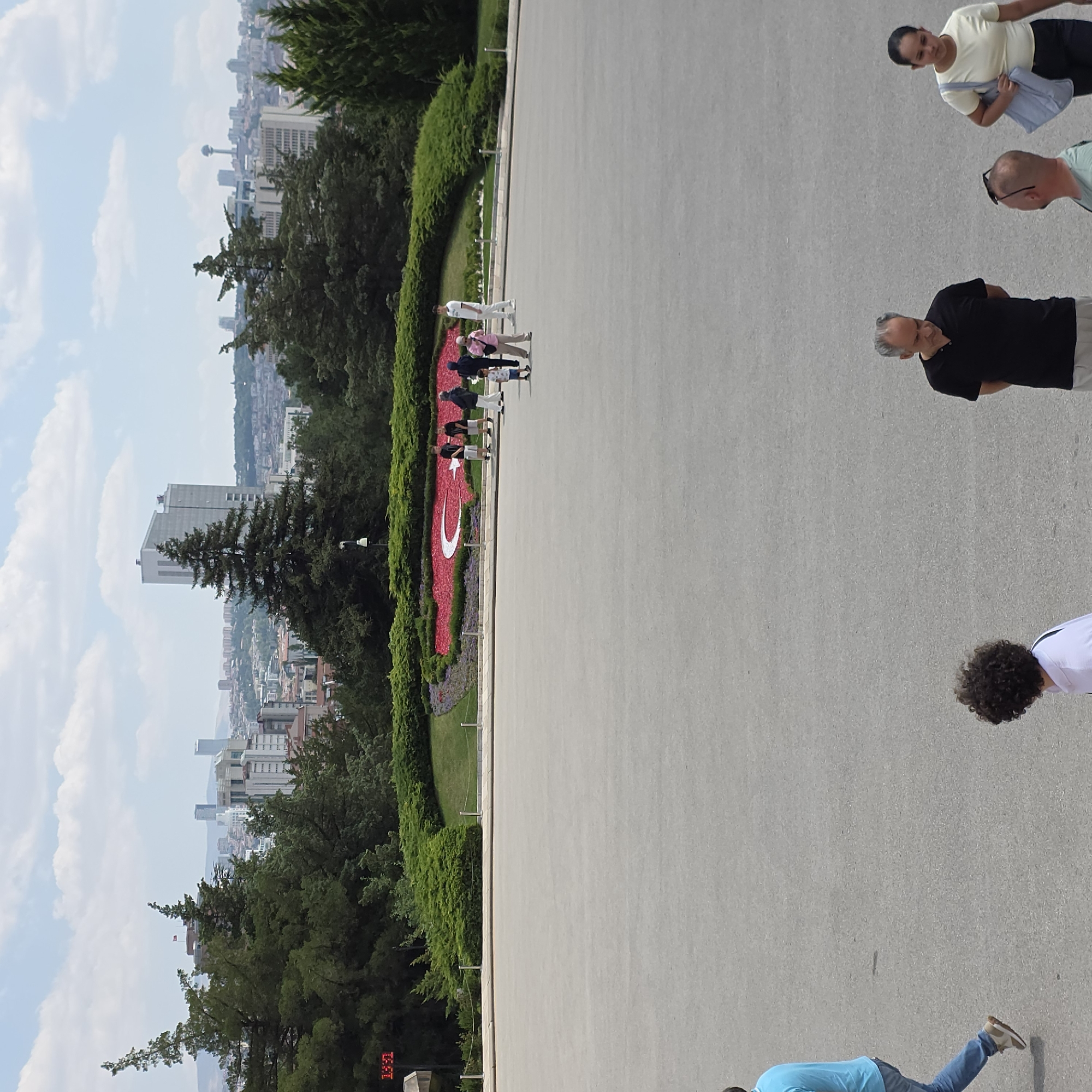 In an outdoor urban environment, a large, flat, grey paved area occupies the foreground. In the mid-ground, a vibrant flowerbed displays a Turkish flag, formed by red and white flowers, against a backdrop of manicured green bushes and trees. Several individuals are observed in this area, some walking and others standing, appearing to be tourists. A digital clock with red numerals displays "13:51". In the background, a panoramic view of a city skyline is visible, featuring numerous buildings of varying heights and architectural styles, including a distinctive tower. The sky is partly cloudy. A person in a blue shirt and jeans is walking on the paved area from the left side of the frame towards the center.