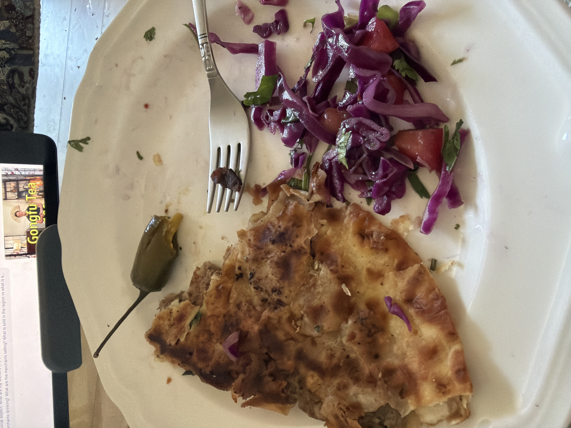 The image shows a close-up, top-down view of a white plate with food and a fork. The plate contains a piece of flatbread, a red cabbage salad with tomatoes and cilantro, and a pickled jalapeño pepper. The flatbread is golden brown and appears to be filled. The salad is a vibrant mix of shredded red cabbage, diced tomatoes, and fresh green herbs. The jalapeño is bright green with a long stem. A silver fork rests on the plate, with a small piece of food on one of its tines.
To the left of the plate, a portion of a smartphone is visible. The screen displays text and an image, with the words "Gongfu Tea" prominently featured in yellow lettering. The text on the phone screen is partially obscured but appears to be descriptive, referencing "merchants drinking," "merchants selling," and "region."
The scene is likely a mealtime setting, given the food on the plate and the presence of a smartphone, suggesting a casual dining experience. The lighting appears to be natural, casting soft shadows. The background is out of focus, but hints of a patterned rug and a white surface can be discerned. No specific time of day or weather conditions can be determined. The overall impression is a close-up shot of a prepared meal, possibly with some informational content being accessed on the smartphone simultaneously.