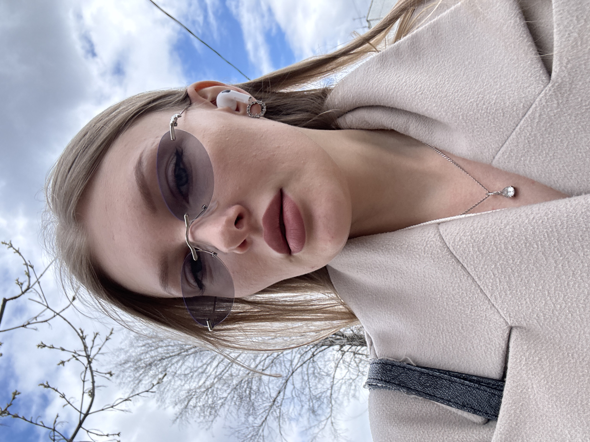 A low-angle, close-up portrait of a woman is captured outdoors in Perm, Russia. Her face is partially visible, with her gaze directed towards the camera, largely obscured by oval, rimless sunglasses with dark, tinted lenses. She has light-colored hair, likely blonde or light brown, swept back from her face. Her lips are full and adorned with a matte, rosy-brown lipstick.
She is dressed in a light-colored, possibly beige or off-white, textured coat, which appears to be structured. Accessories include a white earbud in her right ear, accompanied by a small, silver-toned hoop earring. A delicate silver chain necklace with a small, teardrop-shaped pendant is visible on her collarbone. A detail of a dark, denim-textured strap, possibly from a bag or a coat feature, is seen on her side.
The background features a clear blue sky with scattered white, fluffy clouds. Bare branches of deciduous trees, some showing small, emerging buds, are visible on the left and bottom right, suggesting a late winter or early spring season. A thin, dark utility wire or cable runs across the top right portion of the sky. The scene is illuminated by bright daylight. There is no visible text.