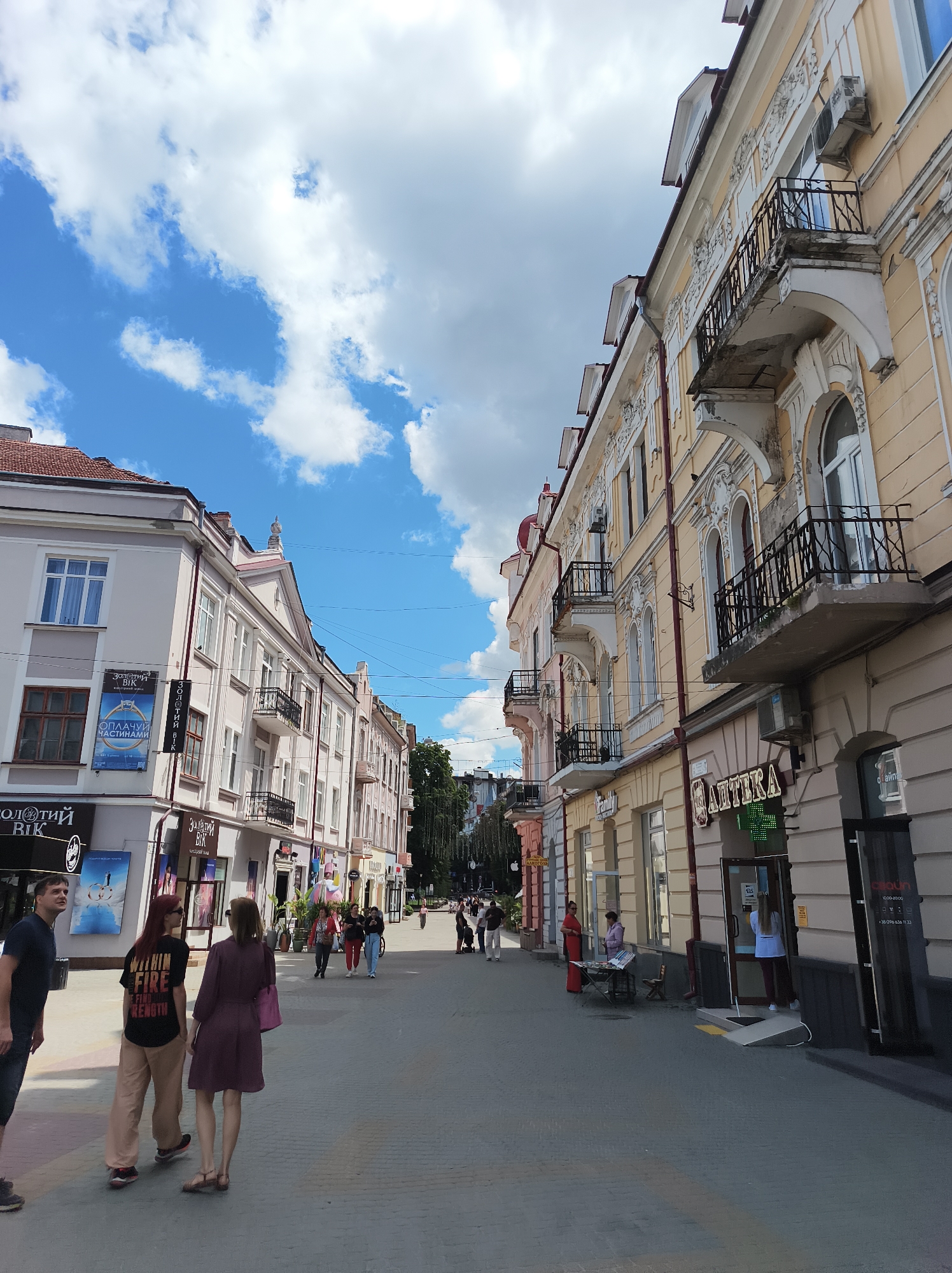 The image captures a street scene in Ternopil, Ukraine, likely on a daytime with a partly cloudy sky. The perspective is from the sidewalk, looking down a relatively wide pedestrianized street flanked by two- and three-story buildings with ornate architecture.
On the left, buildings are painted in pastel colors, with visible shopfronts featuring signs such as "ЗОЛОТИЙ ВІК" (Golden Age) and advertisements for jewelry and watches. Several people are walking or standing on the left sidewalk, including a man in a dark blue t-shirt and jeans looking up, and two women, one in a maroon dress and the other in a black t-shirt with text.
The right side of the street features buildings with a prominent yellow facade, adorned with detailed white stucco moldings, balconies with black wrought-iron railings, and arched windows. Shopfronts on this side include a pharmacy with a neon green sign reading "АПТЕКА" (Pharmacy). A person in a white shirt and dark pants can be seen inside the pharmacy entrance. Further down the street, more pedestrians are visible, as are trees and a few taller buildings in the distance. Streetlights and electrical wires are strung across the street. The street surface appears to be paved with interlocking bricks or stones. The overall impression is of a commercial street in a European city.