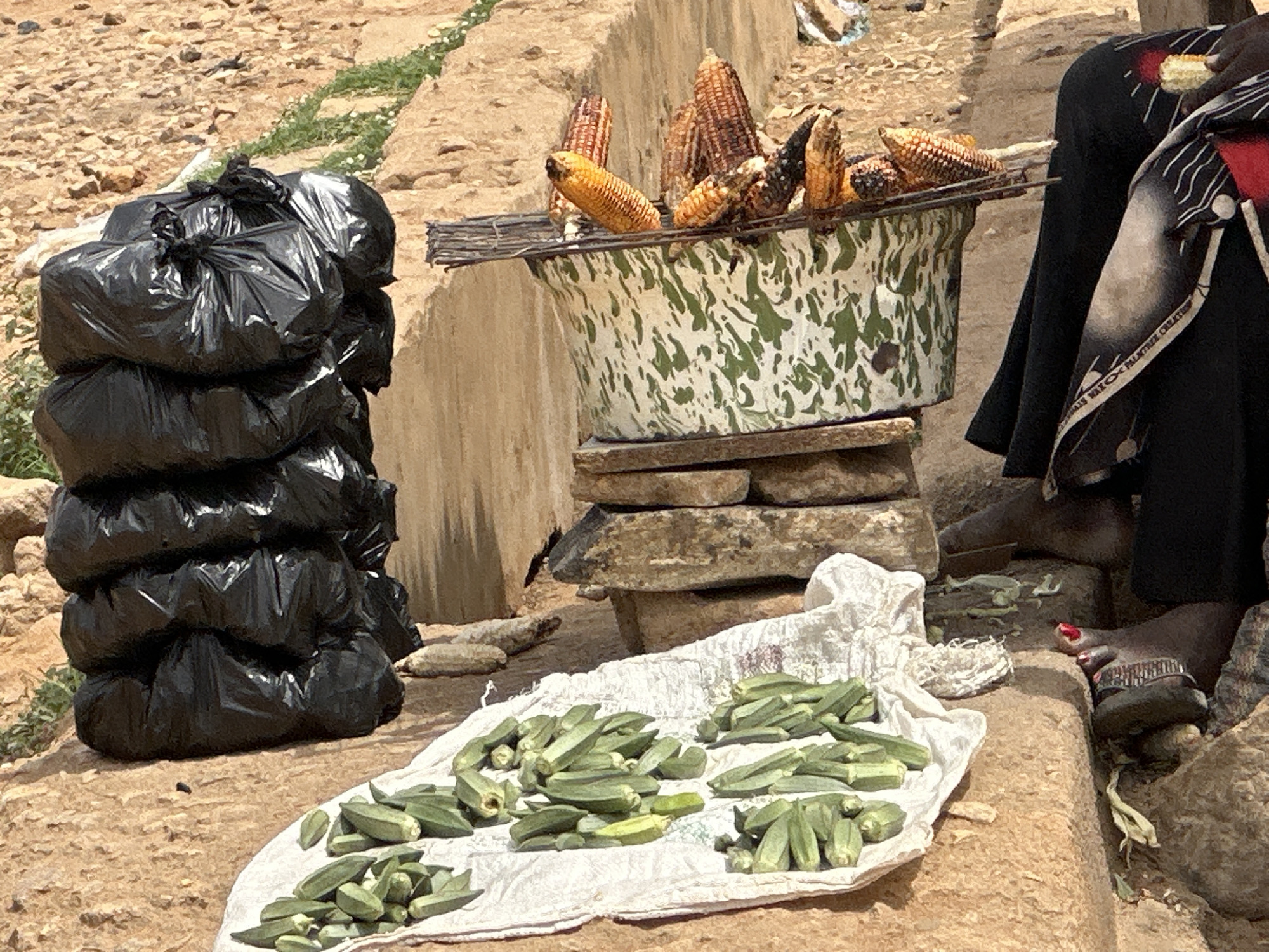 A street market scene in an unidentified location shows a pile of black garbage bags, a bowl of corn on the cob set on stone supports, and a cloth with scattered okra. A person's feet and legs are visible to the right. The setting suggests a rural or less developed area.