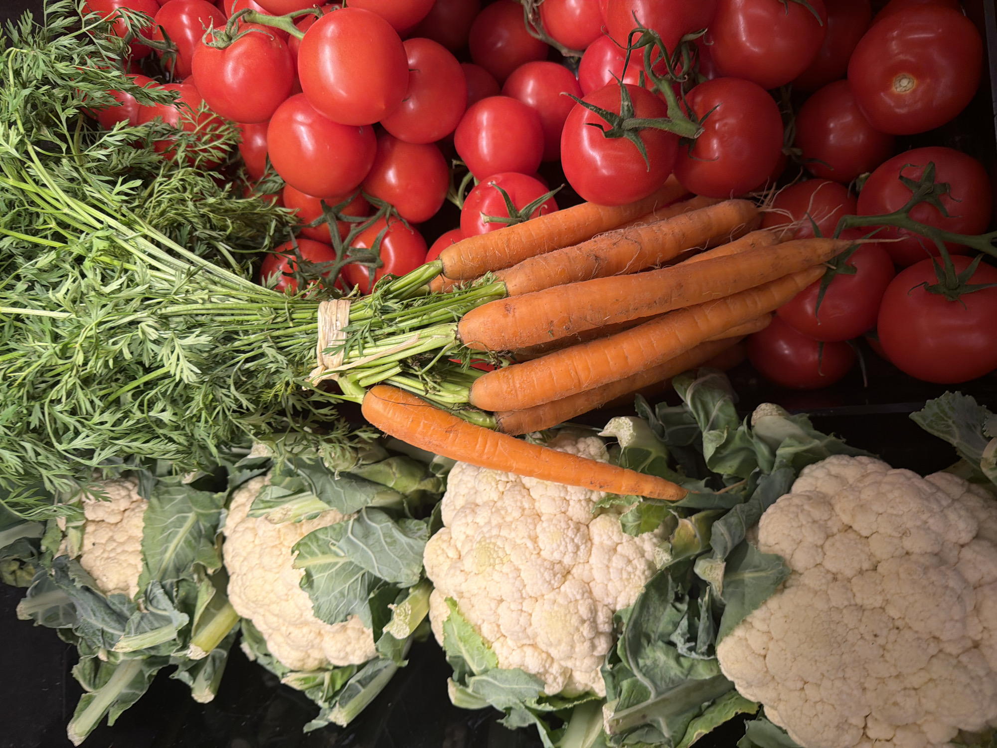 The image presents a vibrant, close-up view of fresh produce, likely displayed in a market setting. The top portion is filled with numerous ripe, red tomatoes, many still attached to their green vines, highlighting their freshness. Below the tomatoes, a neatly bundled bunch of bright orange carrots is prominently featured. Their leafy green tops are clearly visible, tied together with natural twine. The middle ground also shows more tomatoes partially visible amongst the carrots. The bottom section of the frame showcases several large heads of white cauliflower, each surrounded by its deep green outer leaves. The cauliflower appears firm and fresh. The overall arrangement creates a rich palette of natural colors: the deep reds of the tomatoes, the vivid orange of the carrots, and the contrasting greens of the carrot tops and cauliflower leaves against the white florets. The produce is closely packed, suggesting abundance and freshness. Given the context, this could be a display in a market in Paris, France. There are no people, explicit activities, discernible time of day, specific weather conditions, or visible text.