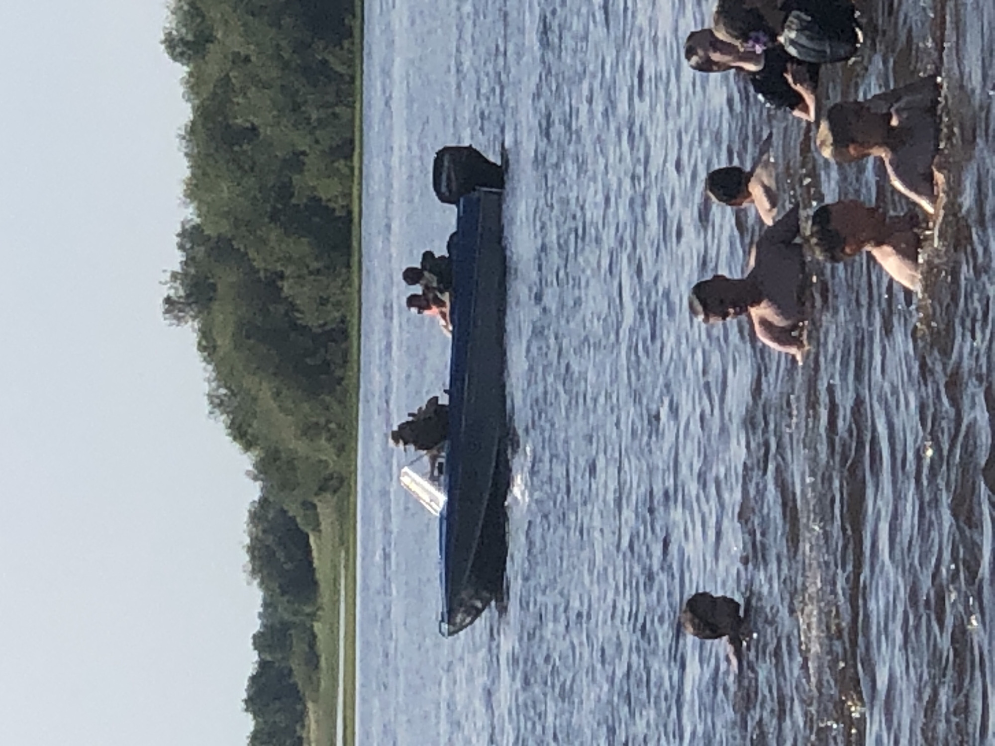 This media file depicts a group of people in a body of water near a wooded shoreline. The scene is dominated by the blue water, which appears to be somewhat choppy. A dark-colored boat with an outboard motor is present in the water, with several individuals inside and around it. Some people are in the boat, while others appear to be swimming or wading nearby.
The lighting suggests it is daytime, likely afternoon, given the bright sunlight casting reflections on the water. The weather appears to be clear and fair. The presence of trees along the edge of the water indicates a natural, possibly rural or semi-rural, setting. The activity suggests recreation or perhaps a boat trip where people are disembarking or swimming.
There are no visible signs of distress or urgency, implying a casual and enjoyable atmosphere. The image's orientation is sideways, making it slightly difficult to discern all details clearly. No text is visible in the image.