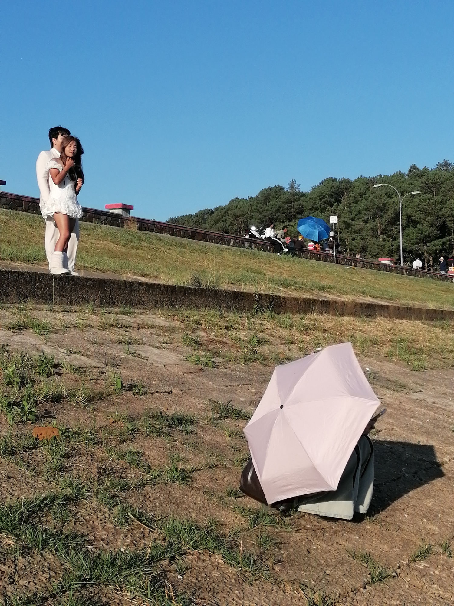 In Da Lat, Vietnam, a couple poses romantically on a raised concrete embankment. The man, wearing a white suit, embraces a woman in a short, white dress. Behind them, a grassy slope leads up to a paved area where several people are seated, some under a blue umbrella, and a street lamp stands tall. In the foreground, a person is seated on the ground under a light pink umbrella, with a dark bag beside them. The sky is clear and blue.
