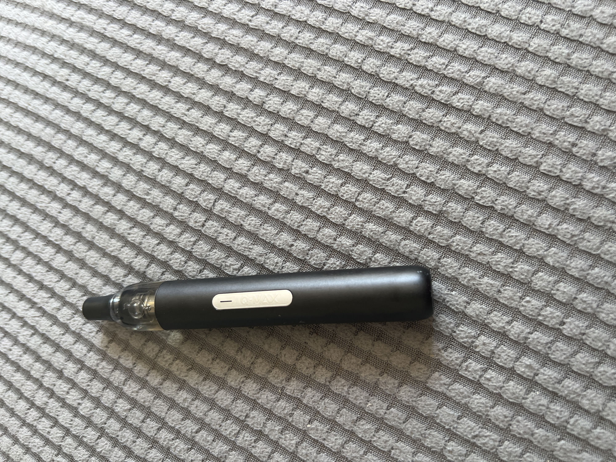 The image shows a black vaping device resting on a gray, textured fabric. The device is cylindrical with a clear tip and a button labeled "Q-MAX". The environment is a close-up view of the fabric's texture, which appears to be a waffle or similar patterned weave. There are no people visible, and no actions are depicted. The location information, Basingstoke and Deane, United Kingdom, is not discernible from the image itself.