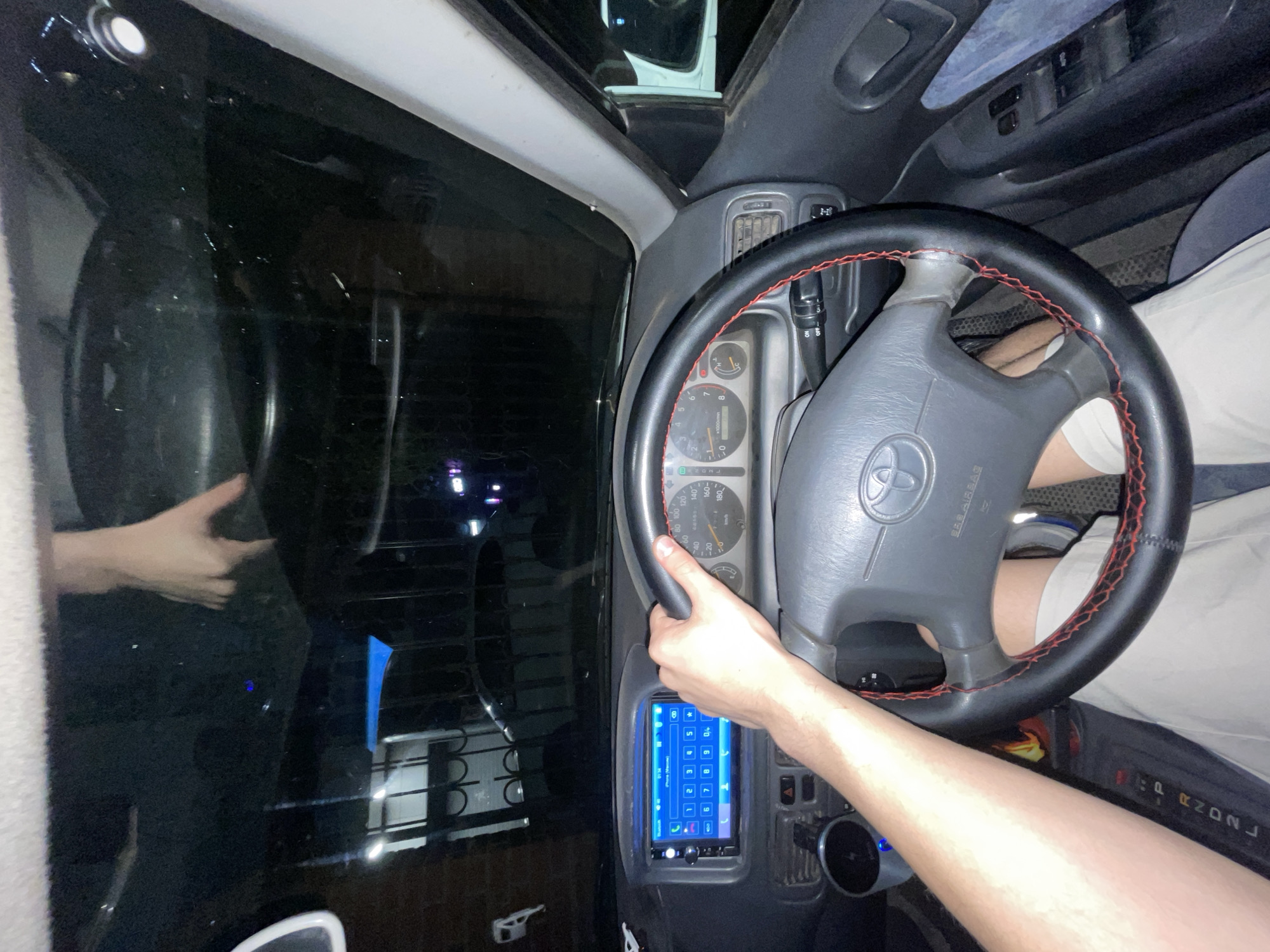 The image is a first-person perspective from inside a car at night. The driver's hands are visible, one on the steering wheel and the other extended upwards, seemingly interacting with the car's windshield or rearview mirror. The steering wheel is black with red stitching and features a Toyota logo. The dashboard shows a digital car stereo with a phone interface and instrument cluster displaying speed and RPM. The surrounding environment is dark, with some lights and reflections visible through the car's windows, suggesting an urban or suburban setting. The presence of a car stereo with a phone interface and the overall interior suggest the vehicle is likely a passenger car. There is no specific visual information within the image that would allow for the identification of the city of Krasnoyarsk, Russia.