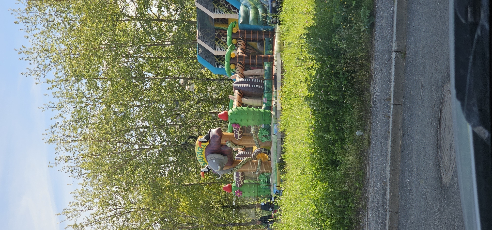 The image shows an inflatable safari-themed playground with an elephant structure and cactuses, set against a backdrop of green trees and a blue sky. There are a few people visible in the background, some standing near the playground. The foreground features a grassy area and a paved road with a manhole cover. The scene appears to be an outdoor public space, likely a park or recreational area, in the city of Novodvinsk, Russia.
