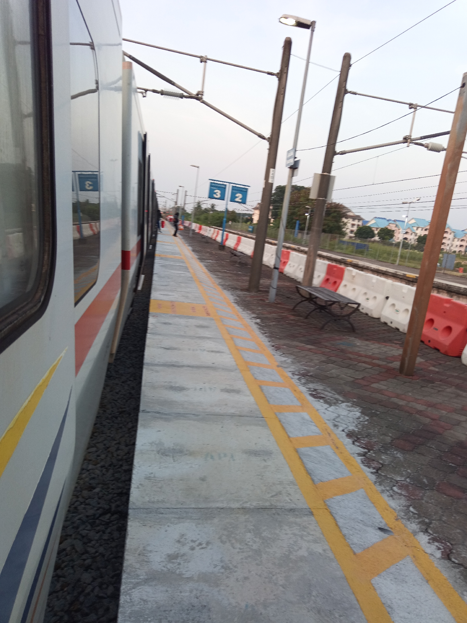 An outdoor, low-angle, full shot shows a train on tracks alongside a platform in Klang City, Malaysia, during the daytime under an overcast sky. The train, which is white with orange and blue stripes, is on the left side of the frame. The platform is made of concrete with yellow markings. Red and white barriers are visible along the edge of the platform, separating it from the tracks. Several streetlights are mounted on poles above the platform. In the background, buildings with blue roofs are visible, along with trees and vegetation. A bench is positioned on the platform near the barriers.