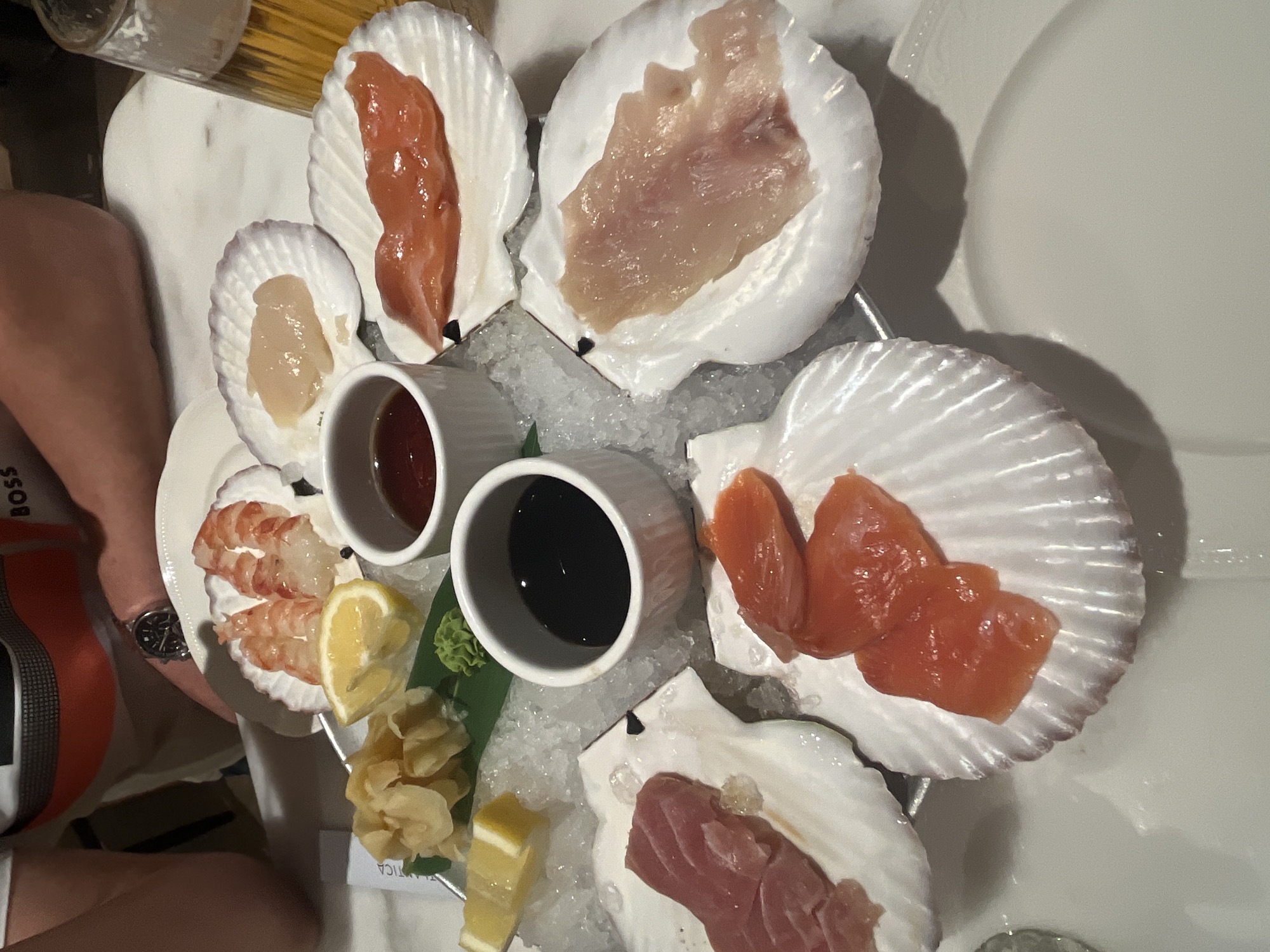 The image shows a raw seafood platter, likely sashimi, presented on a bed of ice in a circular metal tray. The tray contains several scallop shells, each holding different types of thinly sliced raw fish or seafood. There are at least six shells visible, containing what appears to be salmon, tuna, scallops, and possibly shrimp. Two small white ramekins containing dipping sauces, likely soy sauce and a red-colored sauce, are positioned near the center of the tray. A portion of lemon wedges and pickled ginger is also included on the tray. The tray is being held by a person wearing a wristwatch and a shirt with a brand visible on the sleeve. The backdrop includes a white marble surface and a white plate. The location is possibly in Moscow, Russia based on the visible text "Atlantica" and other visual cues.