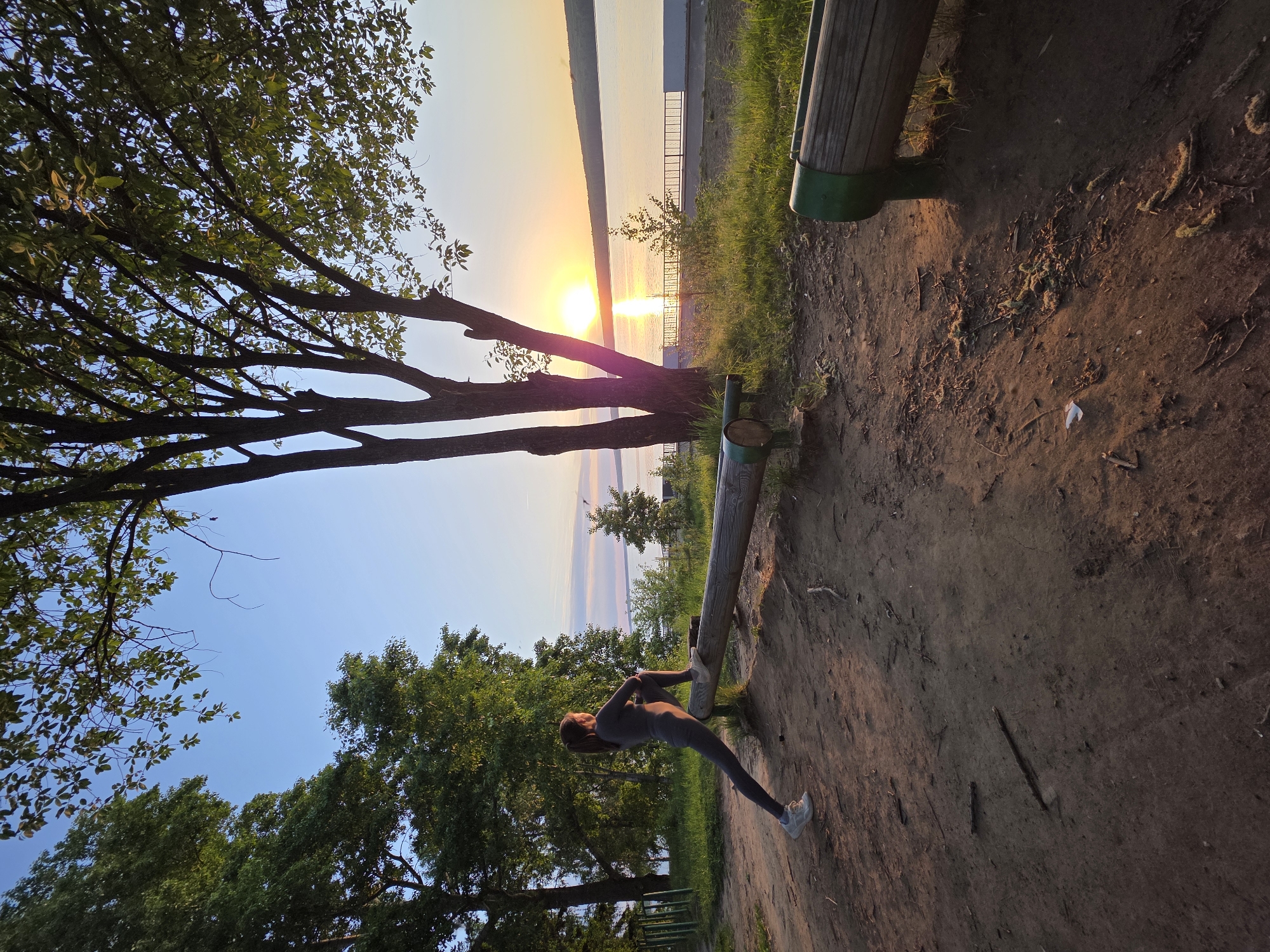 A woman in a grey jumpsuit and sneakers is stretching on a wooden exercise bar in a park in Chaikovsky, Russia. The image is composed with the woman and the park's edge in the foreground, angling upwards to a view of the Kama River at sunset in the background. Trees border both sides of the frame, creating a natural enclosure. The ground is dirt and some vegetation is along the trail.
