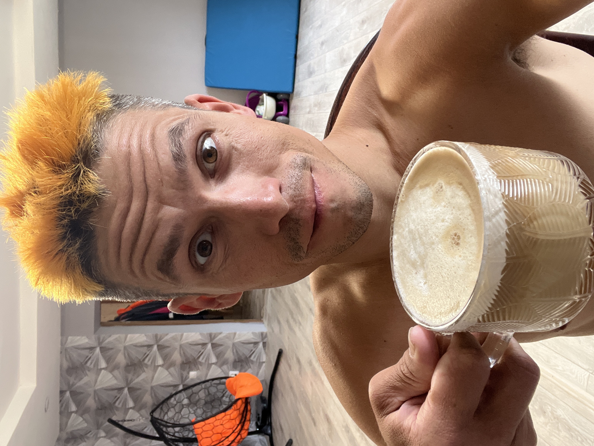 A shirtless man with bright orange spiky hair takes a selfie while holding a clear glass mug filled with a frothy beverage. He is looking directly at the camera with wide eyes and a slight smile. The background shows an indoor setting, possibly a living room or studio, with light-colored flooring. A patterned wallpaper is visible on the left wall, and a large blue mat and a small purple toy are in the background on the right. The man appears to be in his late 20s or early 30s. The scene does not contain any discernible elements that would indicate it is located in the city of Chaikovsky, Russia.