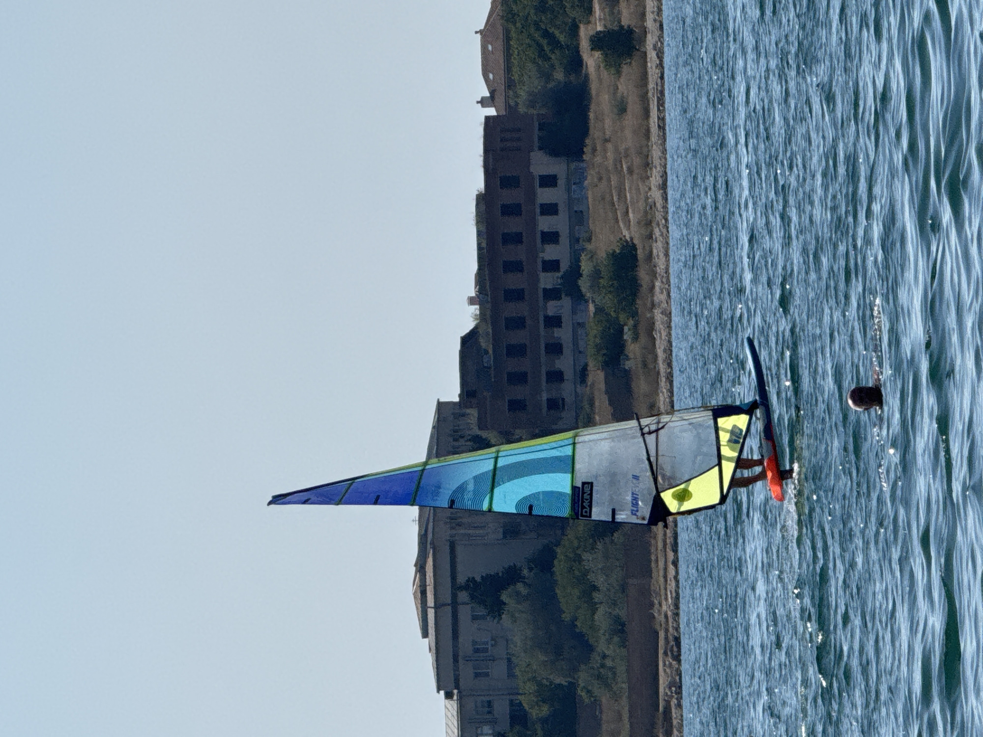 A lone windsurfer maneuvers on the choppy blue waters of Sevastopol Bay, Ukraine, with a backdrop of derelict buildings and lush vegetation lining the shore. The windsurfer's sail is a vibrant gradient of blues and yellows, catching the sunlight as it propels them across the bay. In the foreground, the head and shoulders of a swimmer emerge from the water, their presence adding a sense of human activity to the otherwise serene scene. The buildings in the distance, some appearing to be abandoned or in disrepair, hint at a storied past and provide a stark contrast to the recreational pursuit unfolding on the water. The sky is a clear, pale blue, indicating a pleasant day for outdoor activities.