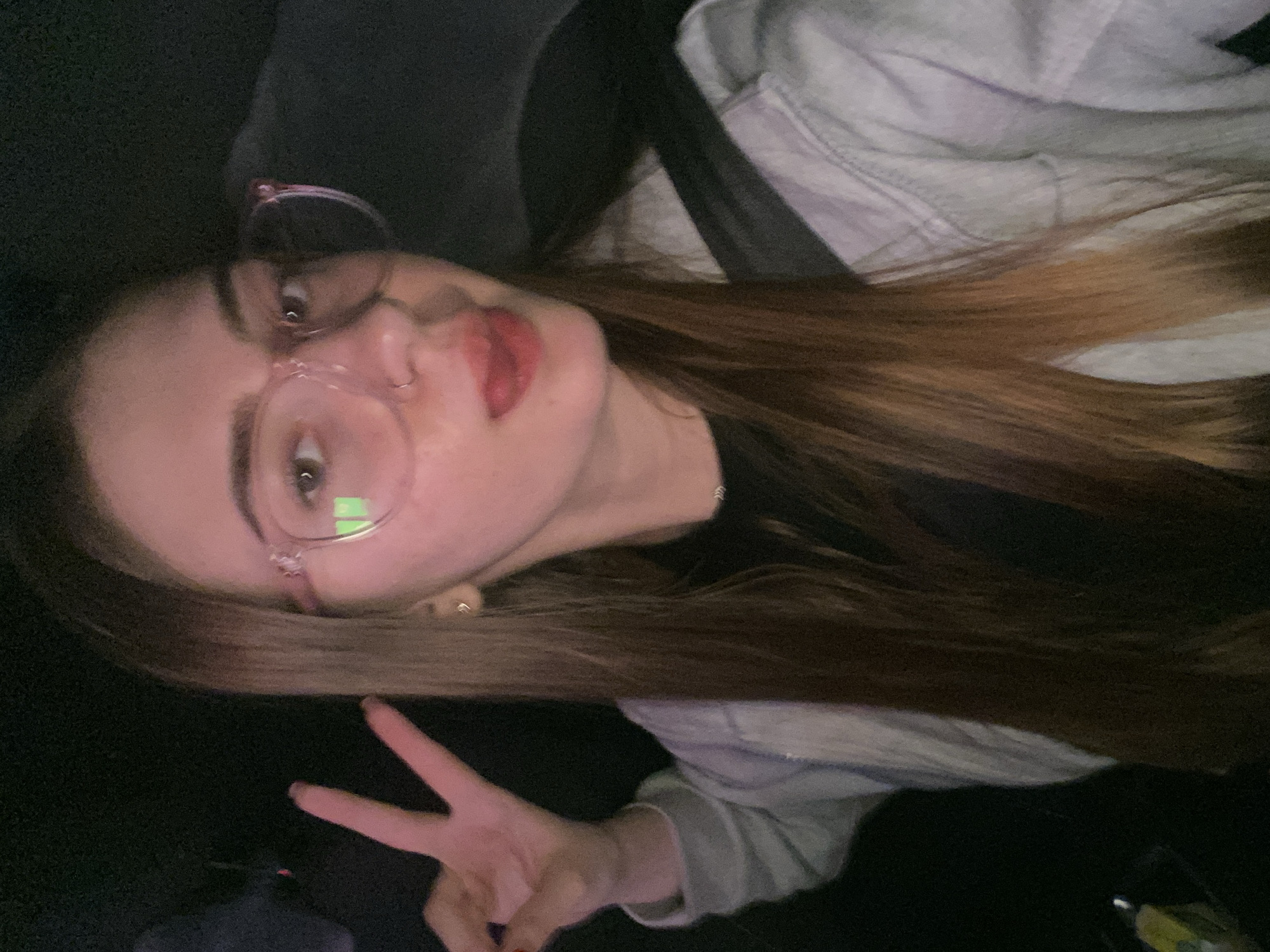 A young woman with long, reddish-brown hair is captured in a close-up selfie. She is wearing large, pink-rimmed glasses and has a nose ring. Her tongue is sticking out to the side, and her right hand is raised, making a peace sign. She is wearing a light-colored denim jacket over a dark top, and a seatbelt is visible across her chest. The background is dark and blurry, suggesting an indoor setting, possibly a vehicle. The image does not provide any specific information or visual cues to associate it with the city of Дьяково, Russia.