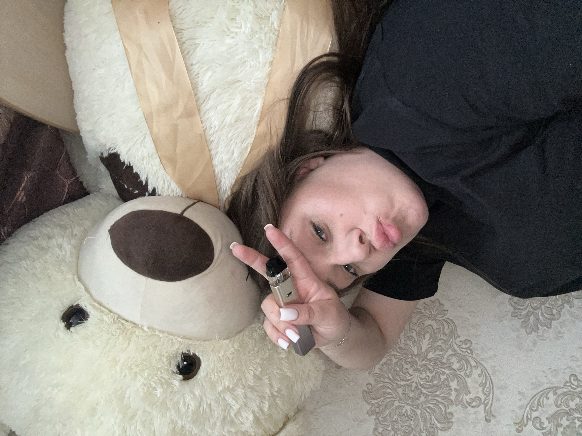 A woman is lying on a floral-patterned surface, likely a bedspread, next to a large, white teddy bear. The bear has brown patches around its eyes and nose. The woman is wearing a black shirt and holding a vaping device in her left hand, making a peace sign with her fingers. Her nails are painted white. The location could be within the city of Chaikovsky, Russia, but that cannot be verified from the image alone.