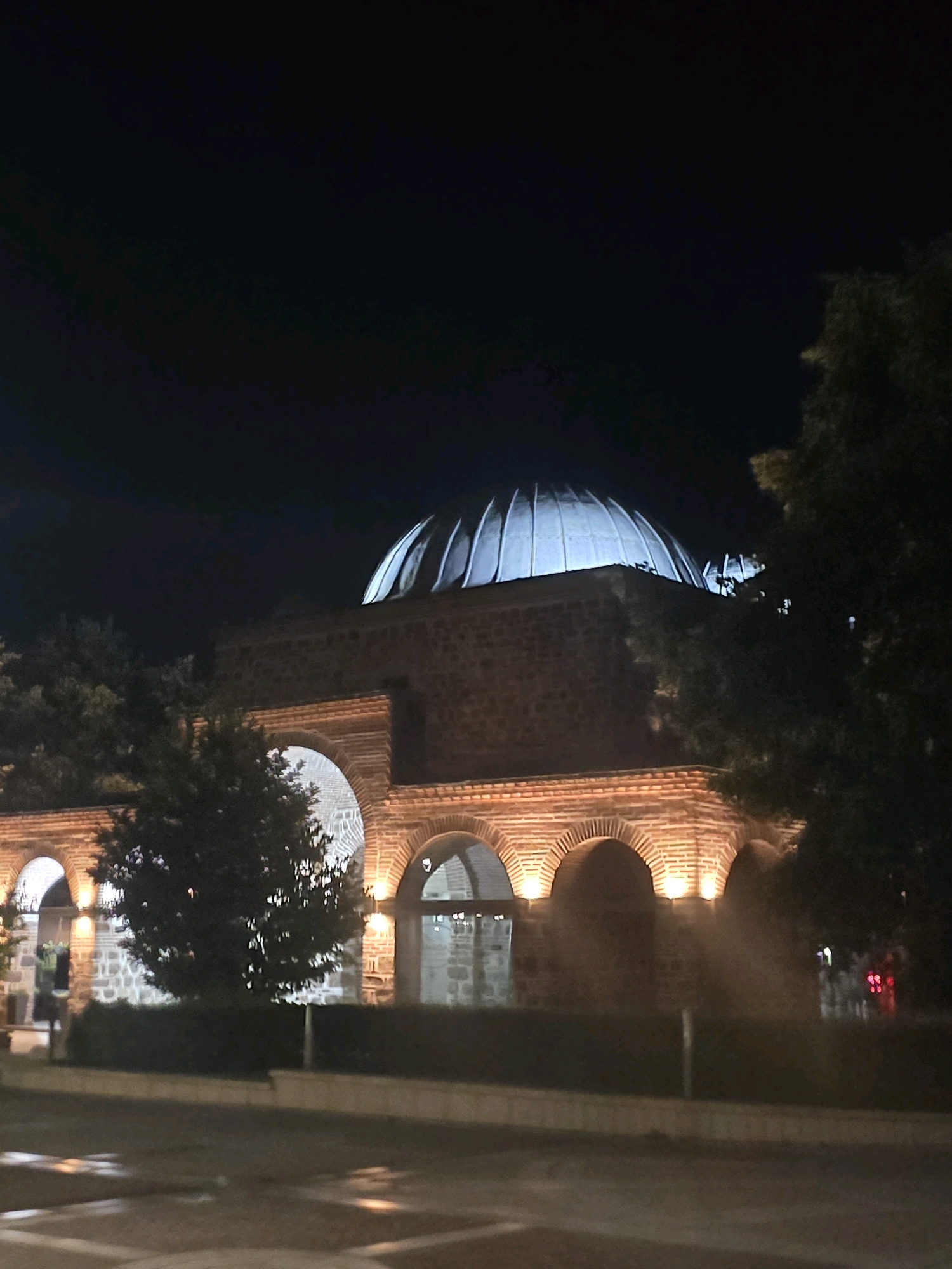 The image shows the exterior of a historic building at night, illuminated by artificial lights. The building features a prominent dome and arched entrances, with stone and brick construction visible. Trees and foliage are present in the foreground and surrounding the structure. The ground in front of the building appears to be a paved surface, possibly a street or walkway, with reflections from the lights. The overall scene is dark, with the night sky forming the backdrop. This building is identified as the former Turkish bath in Yambol, Bulgaria, a historical landmark in the city.