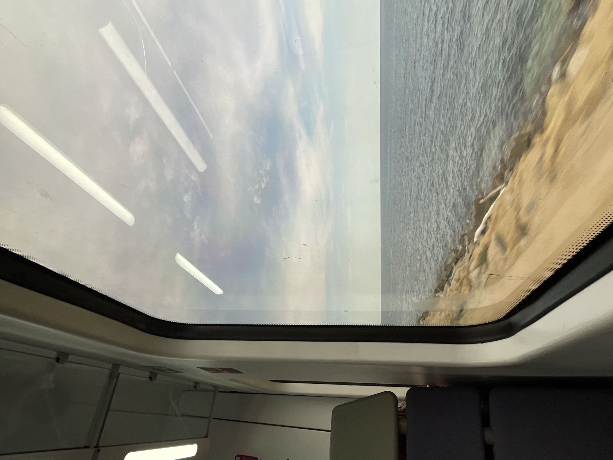 The image shows a view from inside a train compartment, looking out of the window. The window frames a scene of the sea and sky. The sea is a muted blue-grey, with gentle waves. The sky is cloudy, with patches of blue visible. The coastline is visible along the bottom of the frame, with a rocky, sandy shore. The movement of the train is suggested by the slight blur in the foreground landscape. Inside the train, dark blue seats are visible on the left side of the image, and part of a light-colored headrest is also visible. Fluorescent lights are visible on the ceiling of the train compartment.
The coastal train line near Mataró, Spain, offers scenic views of the Mediterranean Sea, with rocky shores and stretches of sand, similar to what is depicted in this image. This particular view would likely be captured on a train traveling along the Maresme coast, between Barcelona and Blanes, which passes through Mataró.