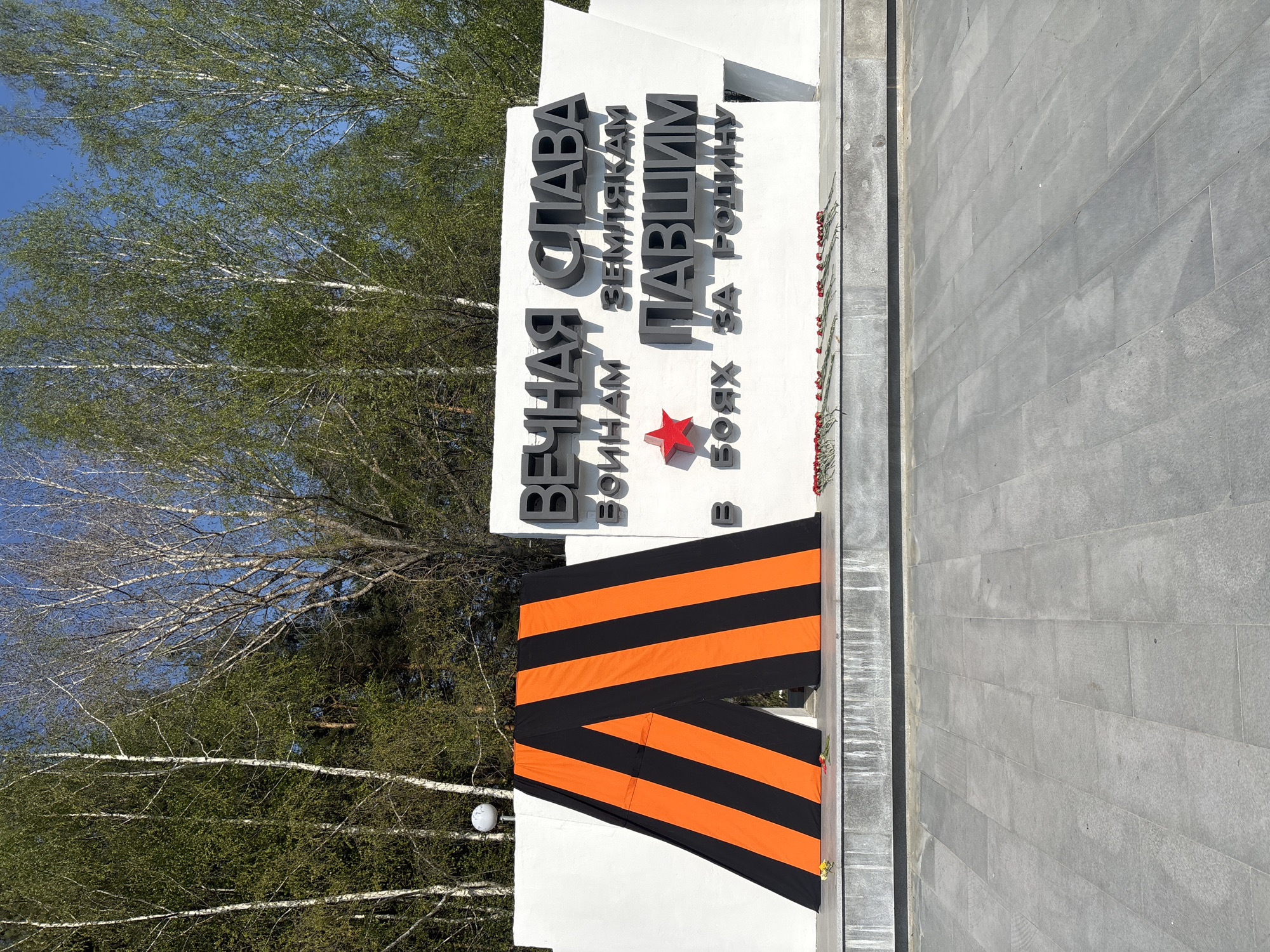 The image shows a memorial in the city of Chaikovsky, Russia. The monument is white with large, dark lettering in Russian: "Вечная слава воинам землякам павшим в боях за Родину," which translates to "Eternal glory to the soldiers, compatriots who fell in battles for the Motherland." A red star is incorporated into the inscription. The monument is draped with a St. George ribbon, a Russian military symbol. Flowers have been laid at the base of the monument. Trees are visible in the background, with a blue sky above. A paved walkway stretches to the right of the monument.
