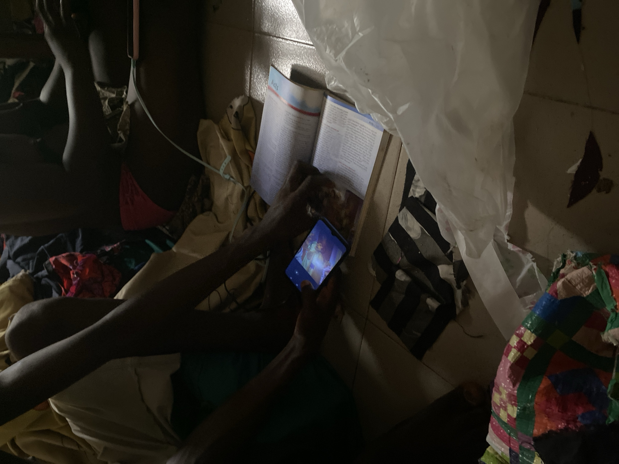 In Jauro Yinu, Nigeria, a person sits on the floor with their legs crossed, holding a smartphone that displays a colorful image. Another person, partially visible, is seated behind them, with a book open in front of them on the floor. The scene is cluttered with various fabrics and a large, white plastic bag. The floor appears to be tiled, and the lighting is dim, suggesting an indoor setting.