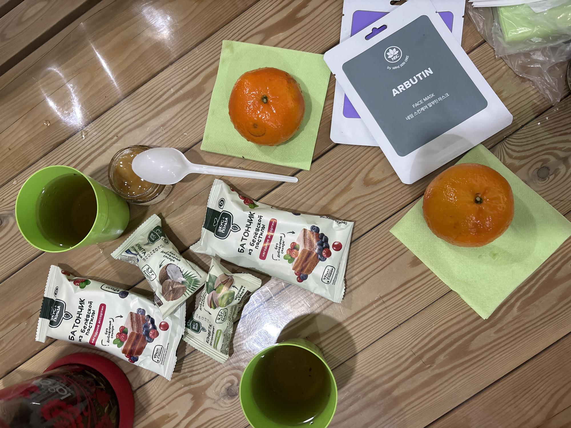 The image captures a top-down view of various items laid out on a light wooden surface, likely a table or floor. The scene suggests a casual snack time or a moment of relaxation in an indoor setting, possibly in Ust-Ilimsk, Russia, given the product labels.
Two vibrant orange mandarins are centrally placed, each resting on a light green paper napkin. Around them, two green plastic cups containing a dark liquid, possibly tea, are visible. One cup is partially obscured by a red base. A small glass jar, holding a light brown, jam-like or honey-like substance, sits with a white plastic spoon resting inside.
Several packaged food items are scattered across the surface. Two larger white packages are prominently displayed, labeled "БАТОНЧИК из белёвской пастилы «Ягодное ассорти»" (Bar from Belev pastila "Berry assortment") from the brand "ВКУС & ПОЛЬЗА" (Taste & Benefit). These labels also indicate "Без добавления сахара" (Without added sugar) and "97 ккал в 1 порции" (97 kcal per serving). Two smaller protein candy packages, one featuring coconut and another pistachios, are also present, labeled "КОНФЕТА протеиновая" (Protein candy) with specific protein content mentioned (e.g., "3,7 г Белка" - 3.7g Protein).
In the upper right corner, several face mask packets are visible, predominantly white and grey. They are labeled "ARBUTIN FACE MASK" in English and "네임 스킨케어 알부틴 마스크" in Korean, under the brand "NSC by NAME SKIN CARE." Some of these masks are inside a clear plastic bag. Reflections of light are noticeable on the polished wooden surface, and a few drops of liquid are visible near one of the green cups. The overall impression is one of a relaxed break, combining light refreshments with self-care products.