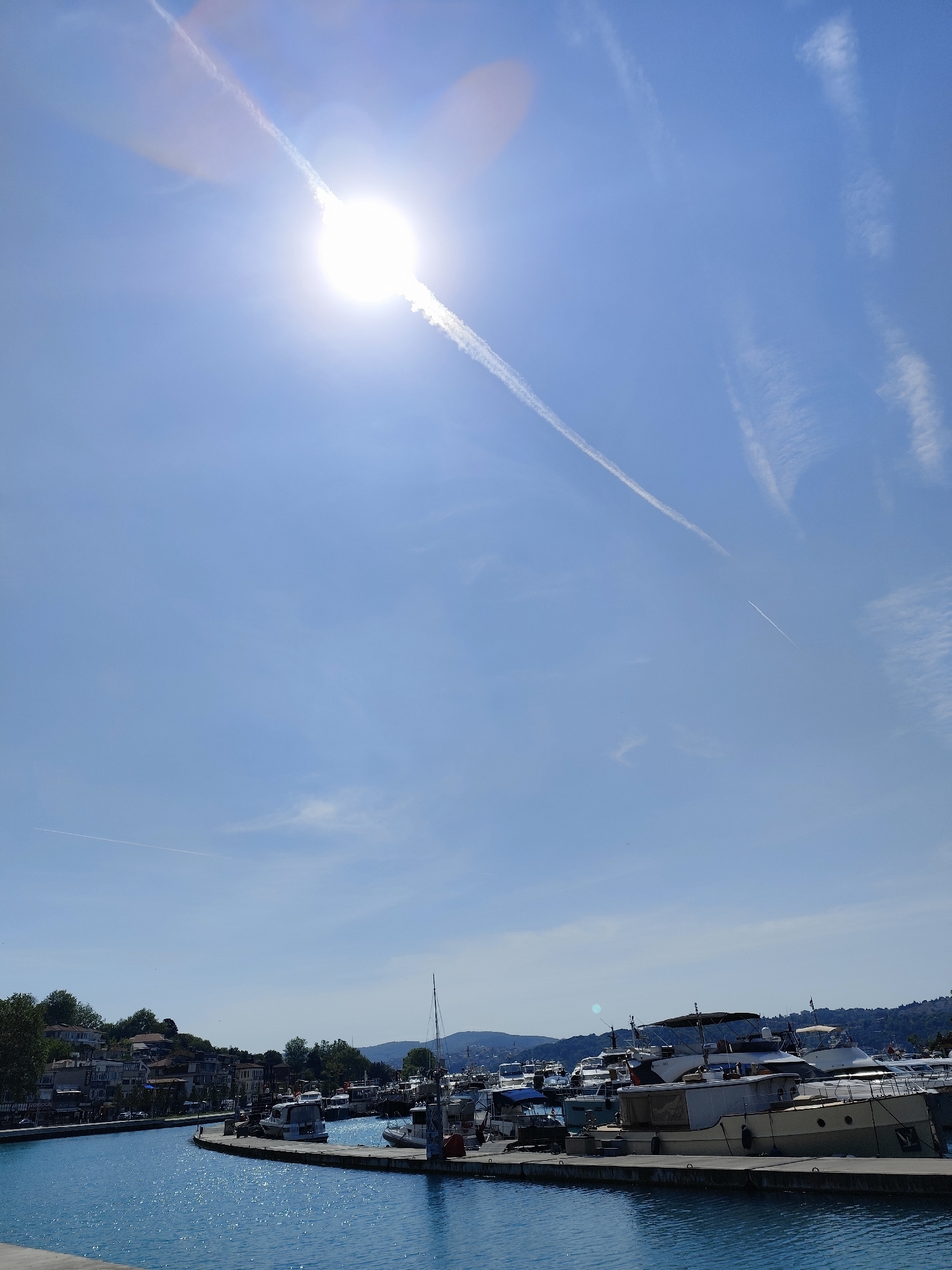 The image depicts a sunny day at a marina in Sarıyer, Turkey. The sky is bright blue with contrails from aircraft visible across the upper portion of the frame. The sun is positioned high in the sky, causing a bright glare. Below the sky, the scene transitions to a marina filled with boats of varying sizes docked along a pier. The water is a deep turquoise color, reflecting the sunlight. On the left side, there's a view of buildings and trees indicating the residential area near the water. In the distance, there are hills or low mountains. No people are distinctly visible.