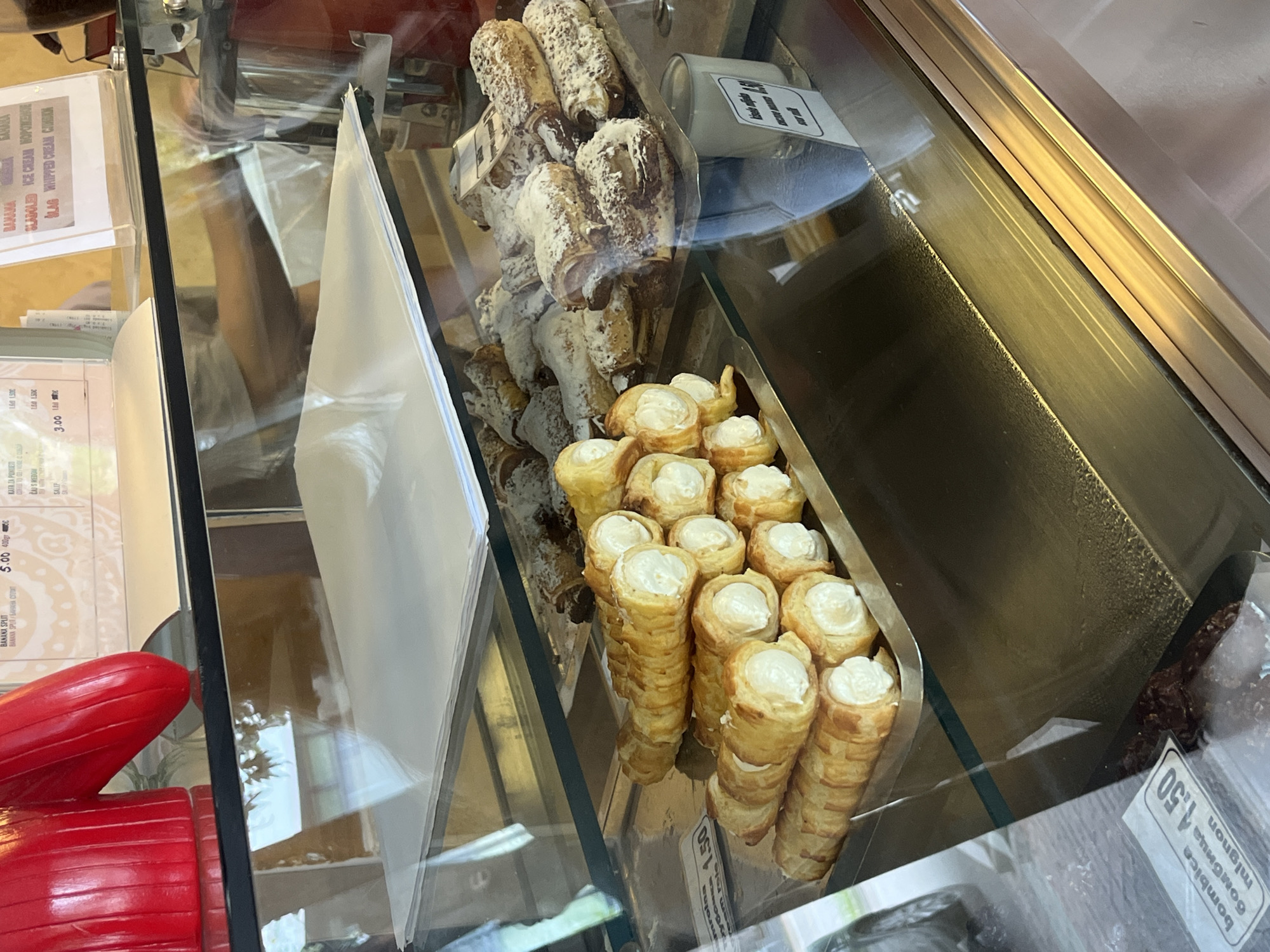The image depicts a pastry display case, likely in a bakery or confectionery in Budva, Montenegro. The case contains several types of pastries, including cream-filled cones arranged neatly in rows and other pastries covered in powdered sugar or nuts, possibly cannoli. Signage is visible, indicating prices in local currency and menu information written in both a local language (Montenegrin) and English. A red, decorative object resembling a hand or glove is visible in the bottom left of the frame. The overall environment is clean and organized, suggesting a professional food service establishment.