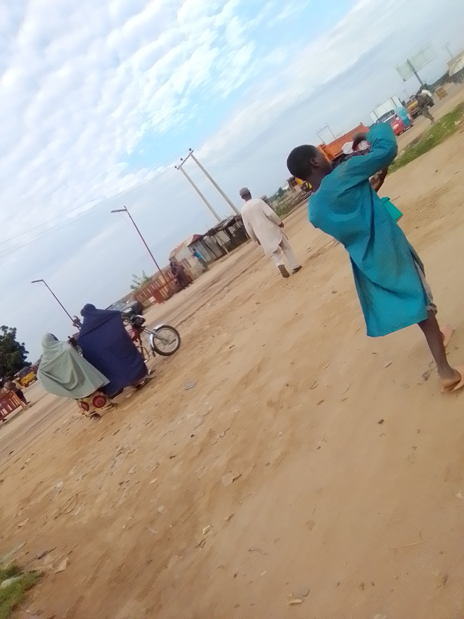 The image depicts a street scene in Gombe, Nigeria, characterized by its dusty, unpaved road. In the foreground, a young boy in a vibrant blue garment is captured from behind, appearing to be walking or standing. His posture suggests he might be looking at something or engaging in an activity not fully visible. Further back, two women are seated on a motorcycle, both covered in traditional Islamic attire, with one wearing a dark blue veil and the other a light grey one. The motorcycle is positioned on the dirt road.
In the mid-ground, a man in a light-colored robe walks away from the camera. Behind him, construction materials and simple structures are visible, hinting at ongoing development or a market area. A bright orange truck is parked on the right side of the road, and other vehicles, including what appears to be a smaller truck or van, are present in the distance. The sky is partly cloudy, with patches of blue visible through the white clouds. The overall environment suggests a typical urban setting in Gombe, with a mix of daily life, transportation, and infrastructure.