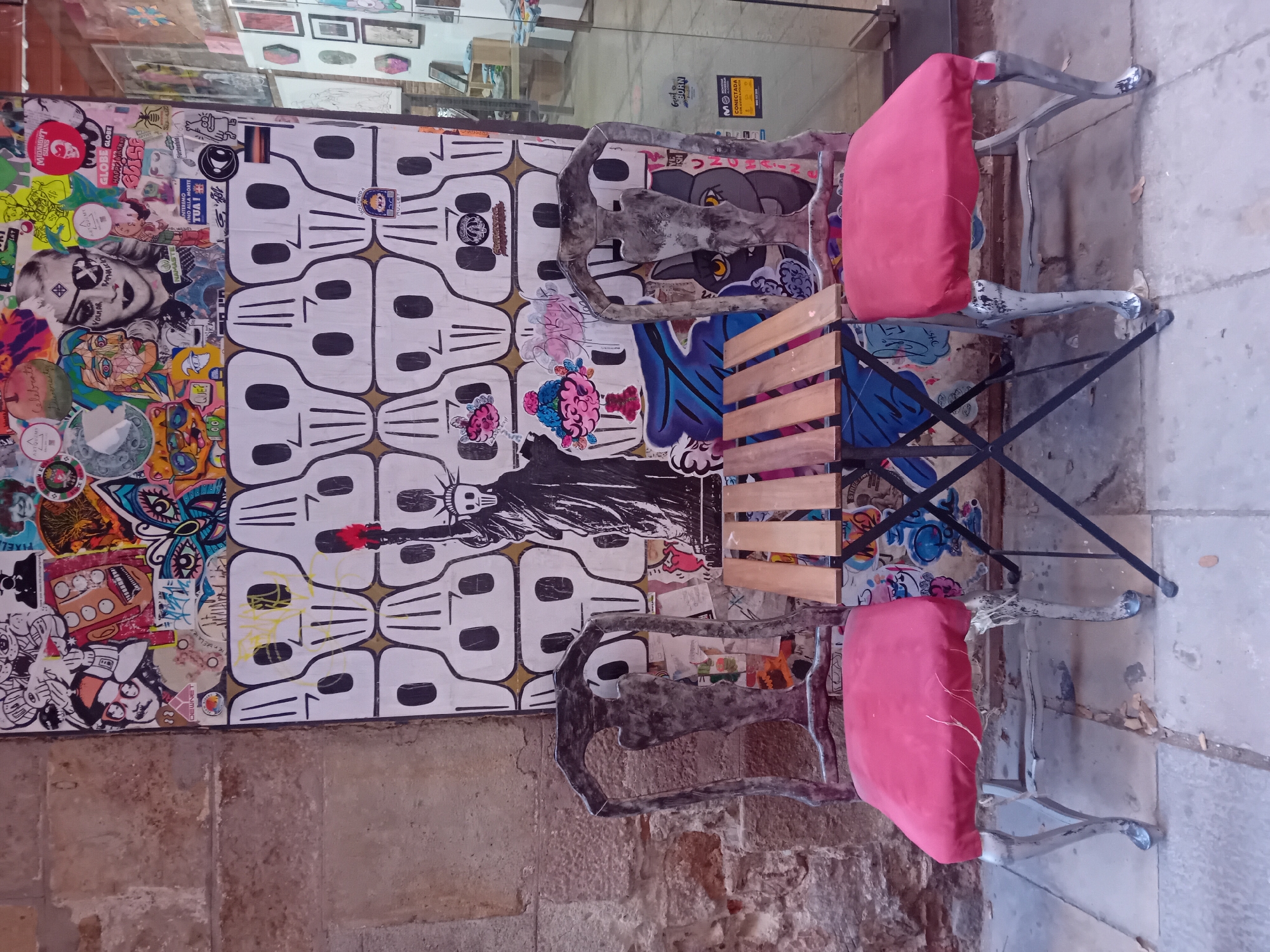 This image captures a vibrant street scene, likely an outdoor cafe or seating area, with a strong focus on urban art and furniture. Two metal chairs with bright red cushions are positioned next to a small wooden table, all resting on what appears to be a stone sidewalk.
The chairs have ornate, somewhat distressed metal frames, giving them a rustic yet artistic feel. Behind the furniture, a prominent wall is covered in a diverse array of street art. The left side of the wall features a dense collage of colorful stickers and graffiti, depicting various characters, symbols, and abstract designs. Notable elements include a large face with sunglasses, cartoonish figures, and bold text. The right side of the wall showcases a patterned graphic of stylized white skulls with black eye sockets and jawlines, arranged in rows. Superimposed on this pattern is a black and white image of the Statue of Liberty, holding what appears to be a torch with a red flame-like element.
In the background, a reflective surface, possibly a shop window, shows blurred glimpses of an interior with framed pictures. The adjacent wall on the left is made of aged stone or brick. The overall atmosphere is eclectic and urban, reflecting a lively public space in Barcelona, Spain.
Visible text from the wall art includes: "PIXEL", "MIDNIGHT", "GLOBE GLOBE", "HAPPY", "VOLMAR", "DEWNET", "NAMASTE NIER", "VINO ALLA MORTE TUA!", "Fürstenfeld", "PAPAYAR", "GORN", "BORN", "MO", "CONECTADA", and "STI".