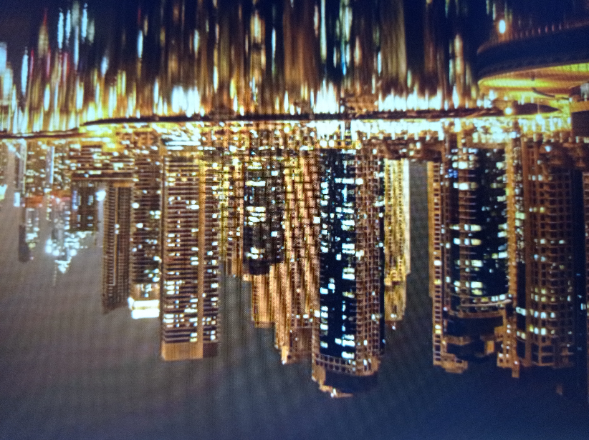 This image depicts a nighttime view of a modern cityscape dominated by numerous illuminated skyscrapers. The buildings exhibit a variety of architectural styles, with many featuring a grid-like pattern of windows that glow with warm, artificial light. In the foreground, a body of water reflects the city lights, creating a shimmering, distorted image of the skyline. The water also shows the silhouette of a boat or a similar vessel. The sky above is dark, suggesting it is either late at night or a heavily overcast sky. The scene is characterized by a high density of tall structures and the vibrant glow of urban illumination against the night sky. There is no discernible indication that this image is related to the city of Mukachevo, Ukraine, as the architectural style and scale of the buildings are more characteristic of major global metropolises.