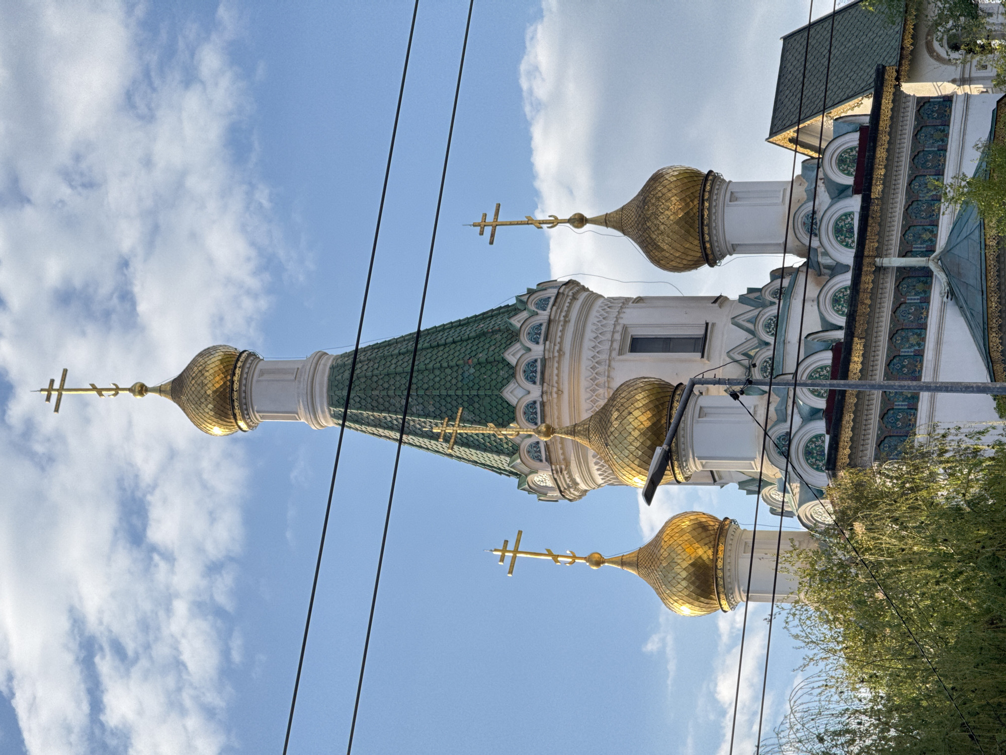 A low-angle shot captures the ornate architecture of an Eastern Orthodox church or cathedral against a bright blue sky with scattered white clouds. The building features multiple gleaming golden, onion-shaped domes, each crowned with a distinctive Orthodox cross. The main roofs are covered in green tiles, creating a striking contrast with the golden domes. The white facade of the building is adorned with intricate decorative patterns in shades of green, blue, and gold, particularly visible around the cylindrical drums supporting the domes and beneath the eaves. Several dark utility wires run diagonally across the sky in the foreground, partially obscuring the upper sections of the church. Green foliage from trees is visible in the lower right corner, adding a natural element to the scene. The clear lighting indicates it is daytime. There is no visible human activity or text in the image. The architectural style is characteristic of Eastern Orthodox churches found in regions like Sofia, Bulgaria, which is provided as the location context.