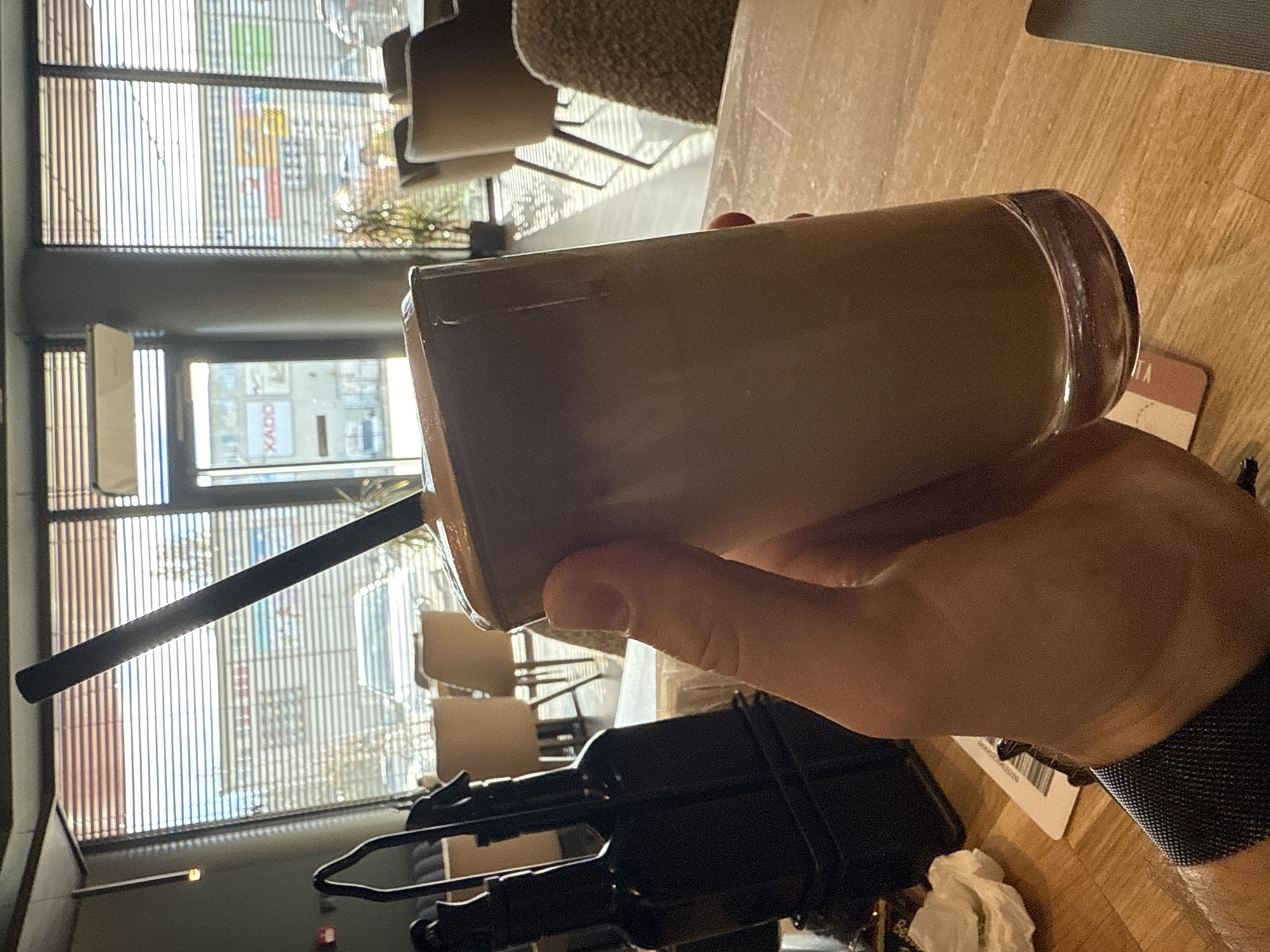 The image depicts a person holding a glass of iced coffee in a cafe or restaurant. The person is wearing a dark-colored wristwatch with a woven band. The drink has a black straw in it. A black object with mechanical-looking parts is on the table below the glass. The table is wooden. A coaster is visible to the right of the glass.
Outside the window, it appears to be daytime, with a street scene visible. The blinds on the window are partially lowered. A store sign with the word "KADO" is seen through the window. Based on the style of the storefront and architecture, the location is likely in Sofia, Bulgaria. There are chairs and tables behind the person, further suggesting a cafe or restaurant setting.