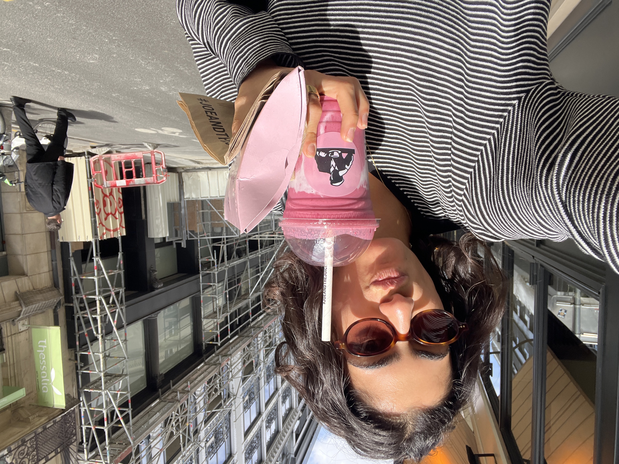 The image, rotated 90 degrees clockwise, presents a close-up of a person holding a beverage and a paper bag on an urban street in Paris, France, under bright daylight.
The main subject is a person, likely female, with dark, wavy hair, wearing dark brown round sunglasses and a black and white horizontally striped long-sleeved top. Her face is visible from the nose down, featuring a slight pout. She holds a large, clear pink cup containing a vibrant berry-colored smoothie with a white layer on top. The cup is adorned with a black and white circular sticker showing a silhouette of a person lifting weights, which is the logo for "Joe & The Juice." A white straw, clearly printed with "#JOEANDTHEJUICE," extends from the drink. She also holds a pink, possibly paper, open container and a brown paper bag, on which the text "#JOEANDTH..." is visible, further indicating the "Joe & The Juice" brand.
The background shows a busy city streetscape. A man in a dark jacket and trousers walks away on a grey pavement, which has faint white markings for a bicycle lane. A small red barrier or cart is also visible on the street level. The buildings lining the street are extensively covered with metal scaffolding, suggesting ongoing construction or renovation. On one building, a distinct green sign with a leaf icon displays the word "Thessalia" in white text. The clear reflections in the building windows indicate a sunny, bright daytime.