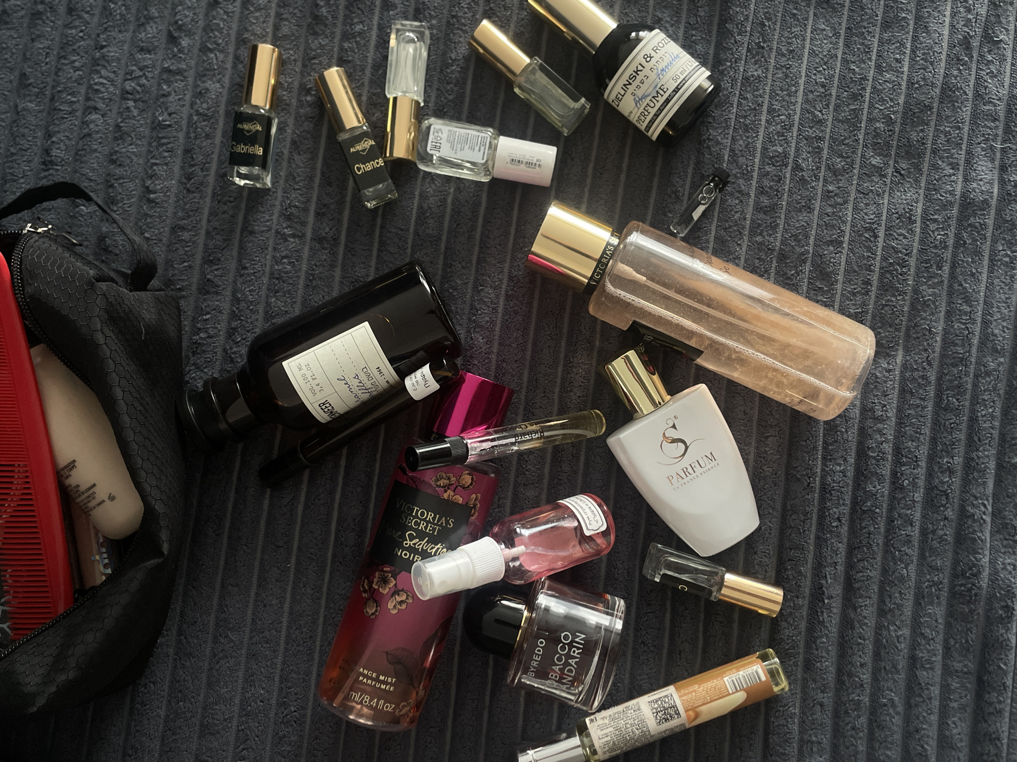 The image shows a collection of perfume and cosmetic items scattered on a dark gray textured surface, possibly a towel or blanket. The items include:
- A black cosmetic bag partially open on the left, containing a red comb and a beige tube.
- Bottles of perfume from brands such as Victoria's Secret (Pure Seduction Noir), Byredo (Tobacco Mandarin), Zielinski & Roze, and other perfumes with labels like "S Parfum la France Essence".
- Smaller sample vials and mini bottles of perfume, some with gold caps labeled "Aurendal Gabriella" and "Aurendal Chance".
- Other cosmetic products such as a spray bottle.
No people are visible in the image. There is no indication in the image of the location being Подшивалово, Russia.