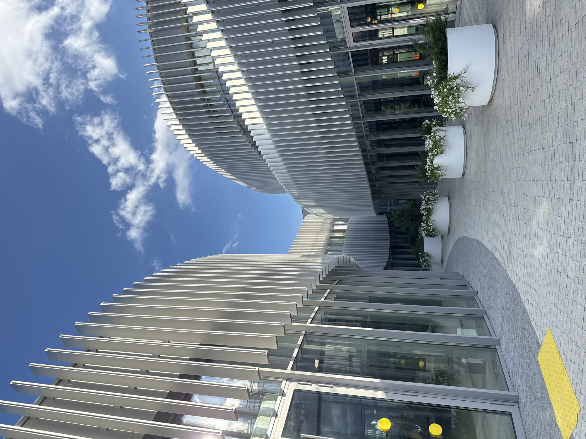 The image shows a modern architectural building in Kazan, Russia, characterized by its curved design and horizontal metal sunshades. The building appears to be commercial or office space, featuring glass facades and rows of white cylindrical planters with flowers and greenery. The ground is paved with light gray stone tiles, and a section of yellow tactile paving is visible. Above, the sky is blue with scattered white clouds.