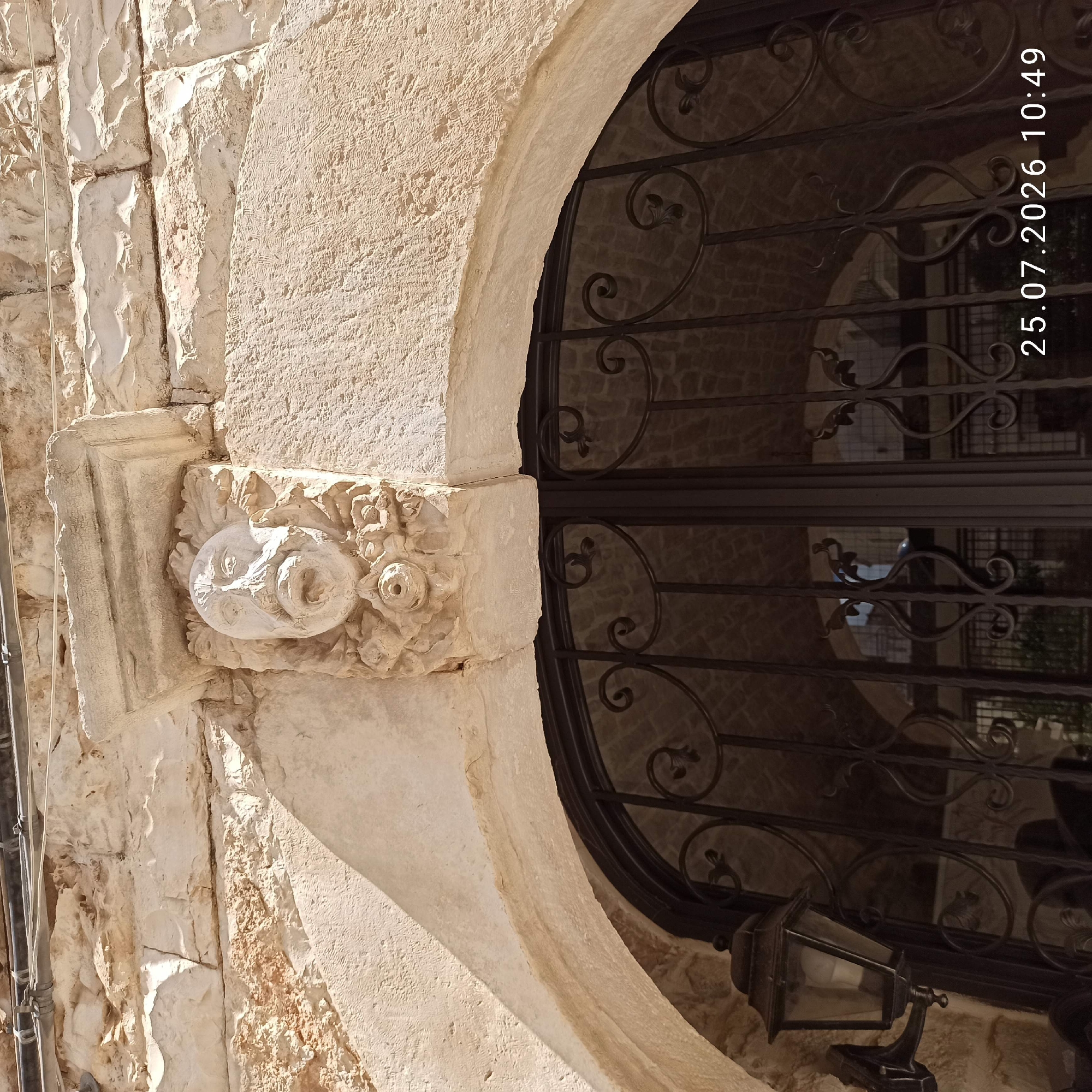 The image captures a detailed view of an architectural facade in Ruvo di Puglia, Italy, specifically focusing on a carved stone face above a wrought-iron arched doorway. The stone face, weathered and textured, is positioned centrally on the facade, surrounded by decorative floral carvings. Above the face, a small stone cornice protrudes. The doorway itself is ornate, featuring intricate black wrought-iron scrollwork, indicative of traditional Italian craftsmanship. To the left of the doorway, a vintage lantern with a black metal frame and glass panes is mounted on the stone wall. The surrounding stonework of the building is rough-hewn and appears aged, with visible layers and textures. The lighting suggests it is daytime, with sunlight casting distinct shadows. The bottom right corner of the image displays a timestamp: "25.07.2026 10:49".