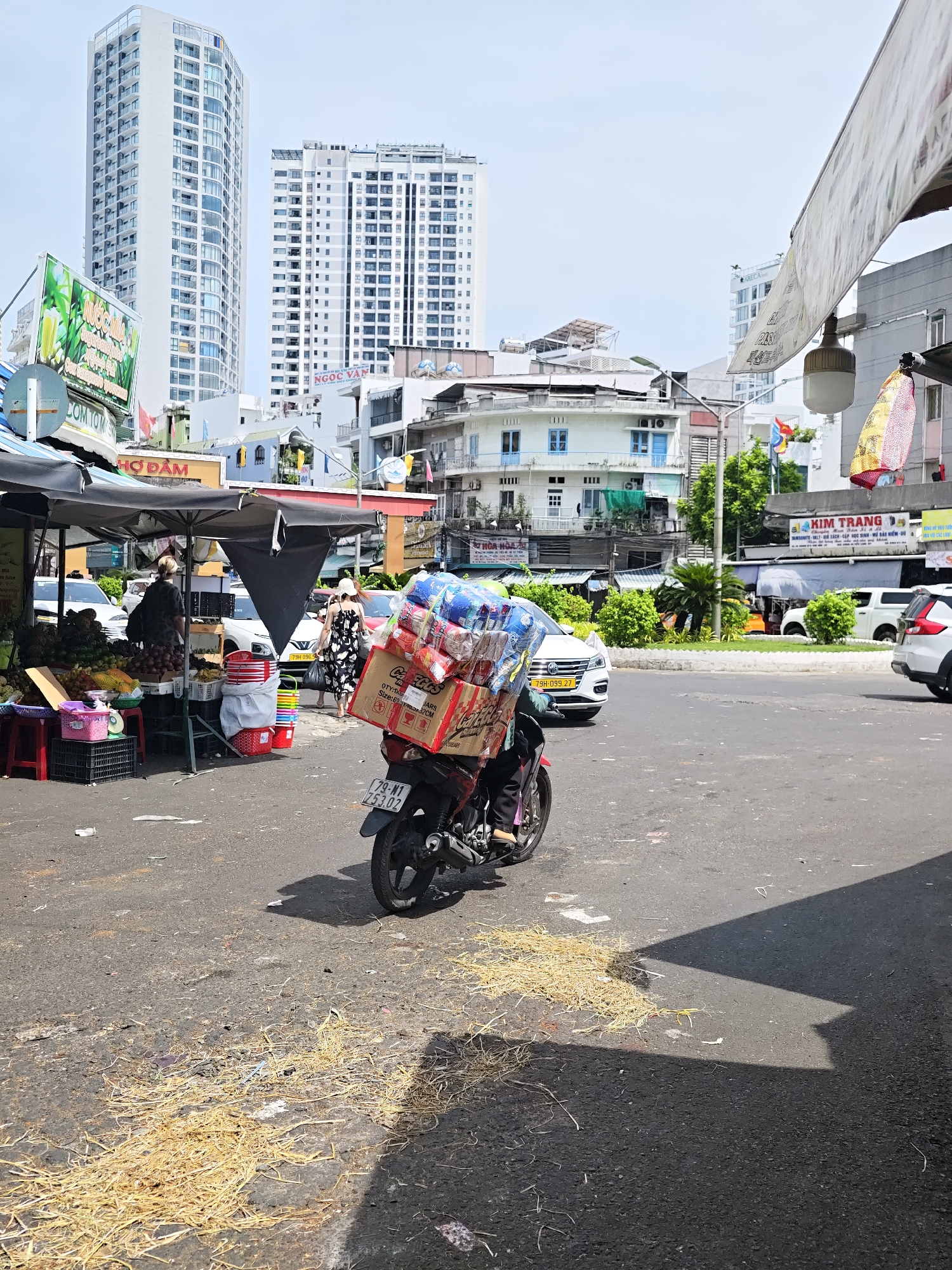 The image captures a street scene in Nha Trang Ward, Vietnam, likely a market area. In the foreground, a motorcycle is parked, loaded with boxes and plastic bottles. A person's legs, wearing black pants, are visible on the motorcycle. The motorcycle has a license plate reading "79-M1 253.02". The street is paved with asphalt and is littered with straw and debris.
To the left of the motorcycle, a market stall is set up under a black canopy. The stall displays a variety of fruits, including pineapples and what appear to be passion fruits. Crates and buckets are also visible at the stall. Two individuals are standing near the stall; one, a woman in a patterned dress and a sun hat, is walking away, while another, a woman in a dark dress and headscarf, is facing the stall.
In the background, several buildings are visible. Two prominent modern high-rise buildings dominate the skyline. Smaller, older buildings with balconies and colorful window shutters are also present. Cars are parked and driving on the street in the mid-ground. Banners and signs with Vietnamese text are displayed on some of the buildings and stalls. The sky is bright and clear, indicating a sunny day. The overall scene depicts a bustling urban environment with elements of commerce and daily life.