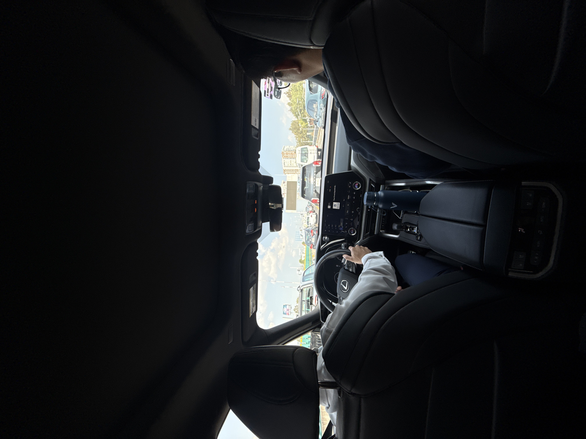 The image captures the interior of a modern car from the perspective of a rear passenger seat, looking forward through the windshield. The car appears to be a Lexus, identified by the logo on the steering wheel.
In the driver's seat, a person wearing a light-colored, long-sleeved shirt is seen with both hands on the steering wheel, indicating they are driving or operating the vehicle. Their head and upper body are partially obscured by the front seat. To the right, in the front passenger seat, another person is partially visible, primarily their head, wearing glasses and with dark hair.
The car's interior features dark leather seating and dashboard elements. A center console is visible, equipped with a large infotainment screen and climate controls. A dark blue water bottle, featuring a white "A" logo, is placed in a cup holder between the front seats.
Through the windshield, an exterior scene unfolds, showing an urban environment with multiple vehicles, suggesting the car is in traffic or stopped on a street or highway. The sky is bright with scattered clouds, indicating it is daytime. The overall impression is one of a routine car journey in an urban setting, likely Mexico City as per the provided context. No specific location cues like landmarks are distinctly visible in the traffic scene itself, beyond the general urban setting.