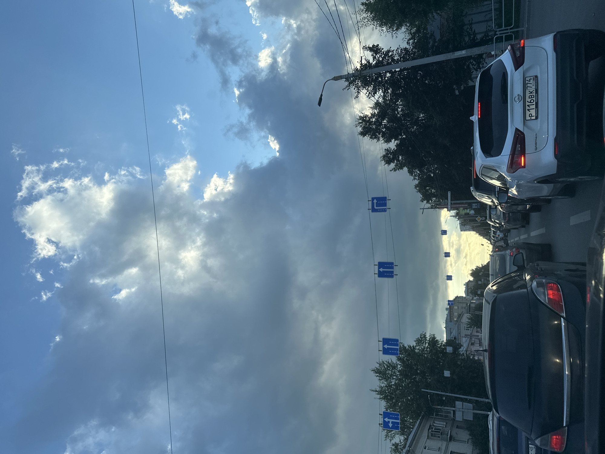 A vertical, eye-level, medium shot captures a traffic jam on a cloudy day in an urban setting. The sky above is a mix of dark, heavy clouds and patches of bright blue with sunlight breaking through, creating dramatic lighting. Several blue traffic signs with white arrows indicating lane direction are suspended over the road. The foreground is dominated by the rear of a black hatchback car. To its right, a white Nissan crossover SUV is visible, with its license plate displaying "P 116BK 774". Multiple lanes of traffic are backed up, with cars stretching into the distance. On the left side of the road, buildings with multiple stories are visible, and on the right, trees and a lamppost are present. The overall impression is of a stalled commute under a dynamic sky.