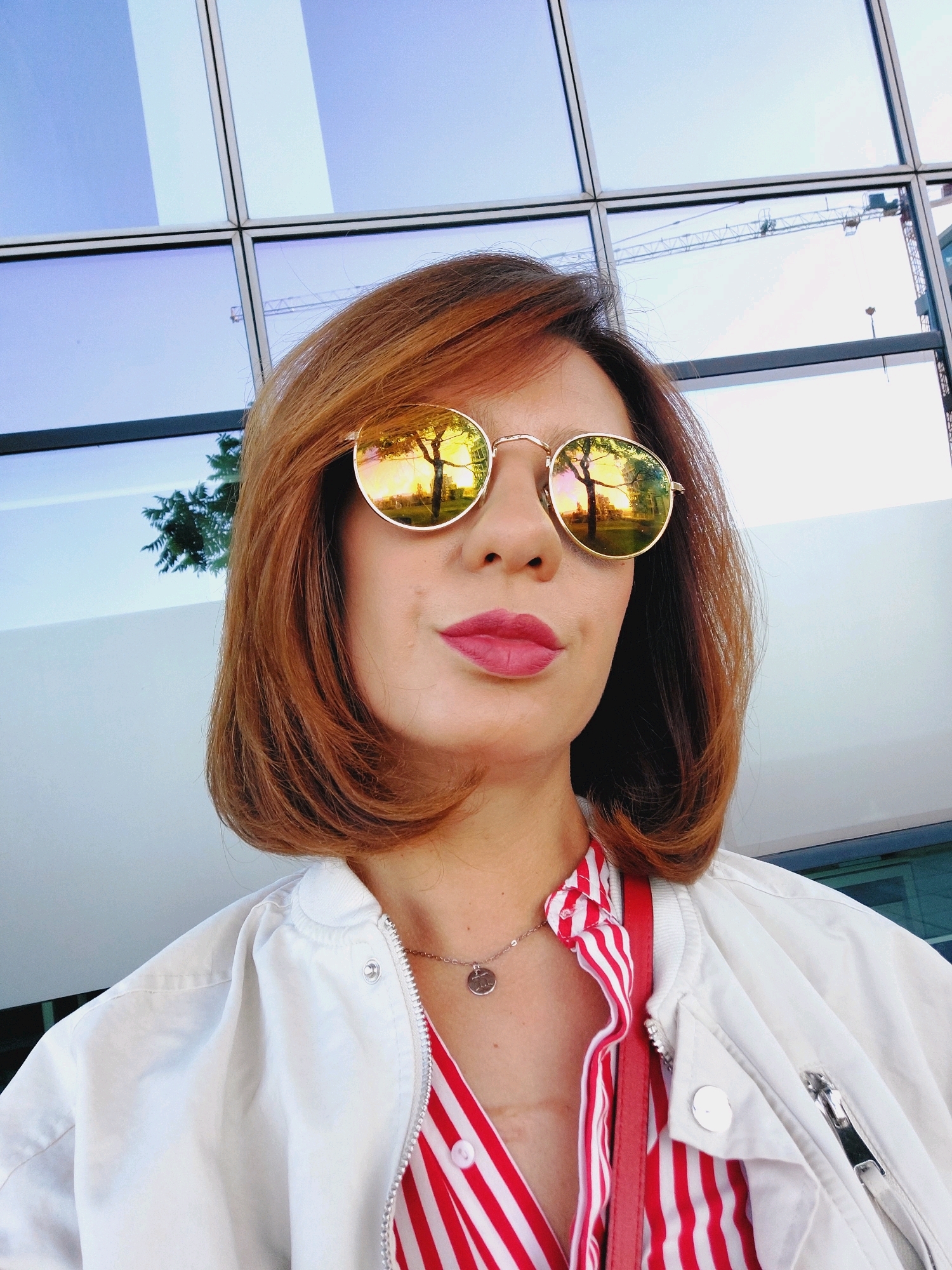 A medium shot shows a woman with auburn bobbed hair and red lipstick wearing round, gold-rimmed sunglasses, a white bomber jacket over a red and white striped shirt, and a silver pendant necklace. Her sunglasses reflect a tree with yellow leaves and a building. In the background, a glass building with large windows displays a reflection of a construction crane. The image is likely taken outdoors in an urban setting, possibly Sofia, Bulgaria, given the architecture.