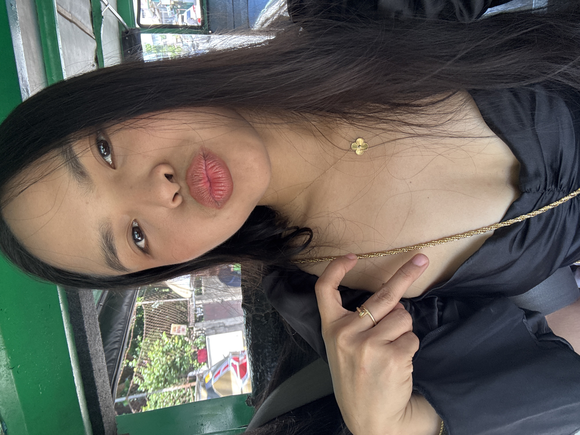 The image is a close-up selfie of a person, rotated 90 degrees counter-clockwise, showing their upper body and head. The subject has long, dark hair flowing over their shoulder and chest. They are looking towards the camera with a playful expression, featuring pursed lips in a "kissy face" pose. Their dark eyes and prominent eyebrows are clearly visible.
The person is wearing a black top or dress with a gathered neckline. Two gold-colored necklaces are visible: a delicate chain with a small four-leaf clover pendant, and a thicker, braided chain draped across the chest. The person's right hand is visible in the lower right, with a simple gold-colored ring on the ring finger.
The background reveals a green metal frame or structure on the left side. Reflections in what appears to be a window or mirror show an outdoor scene with green foliage, trees, and indistinct buildings or infrastructure, suggesting the photo was taken inside a vehicle or structure overlooking an exterior view. The lighting indicates it is daytime. No legible text is visible. The scene is located in Davao City, Philippines.