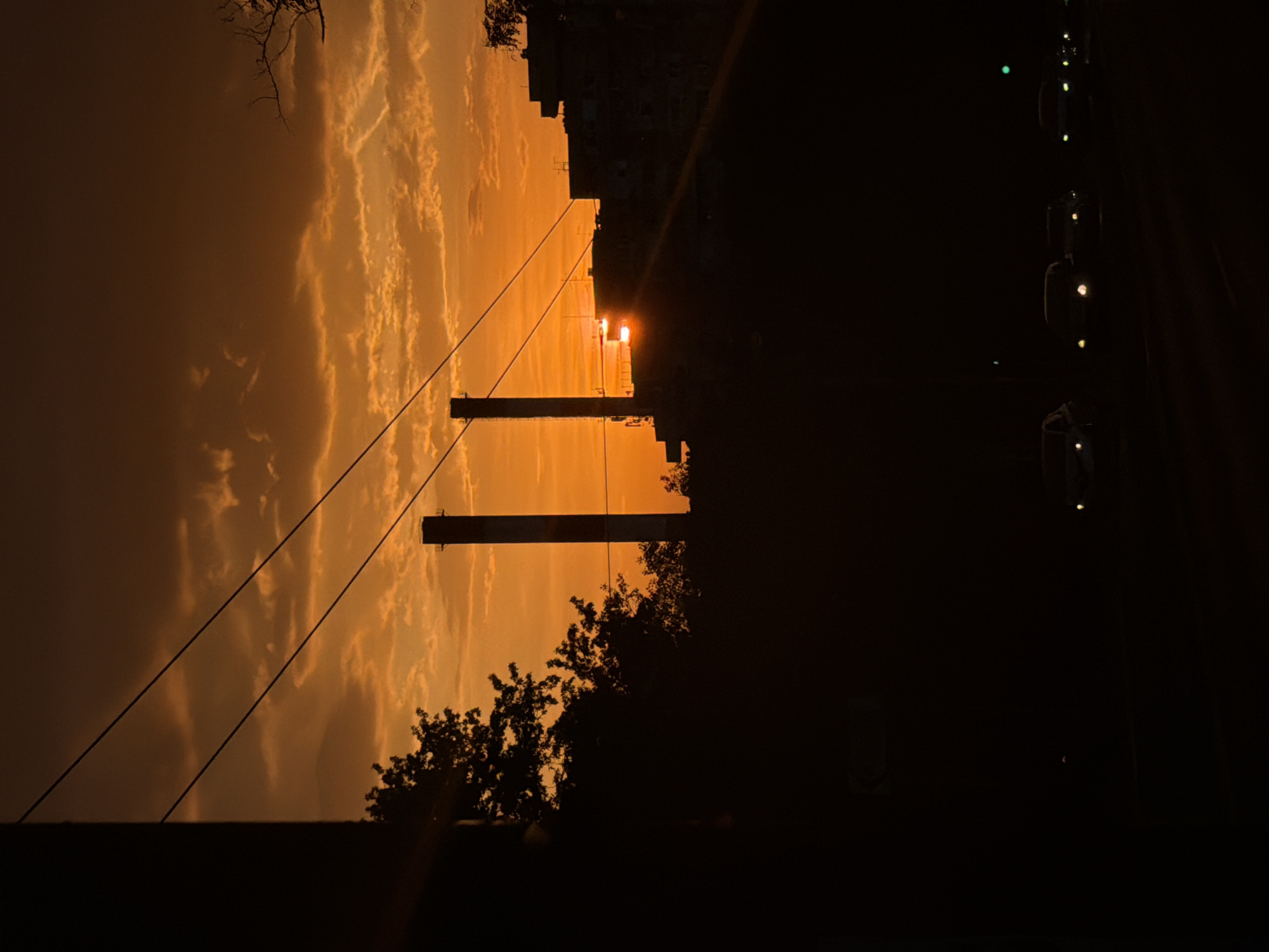 The image depicts a twilight cityscape in Vozdovac Urban Municipality, Serbia. In the foreground, silhouetted trees and rooftops frame an orange-hued sky filled with clouds. Two large, dark industrial structures stand in the center of the image. Overhead power lines run diagonally across the sky. On the right side, a dark road with several points of light, likely from car headlights, is visible. The overall scene captures a moment of urban sunset or sunrise.